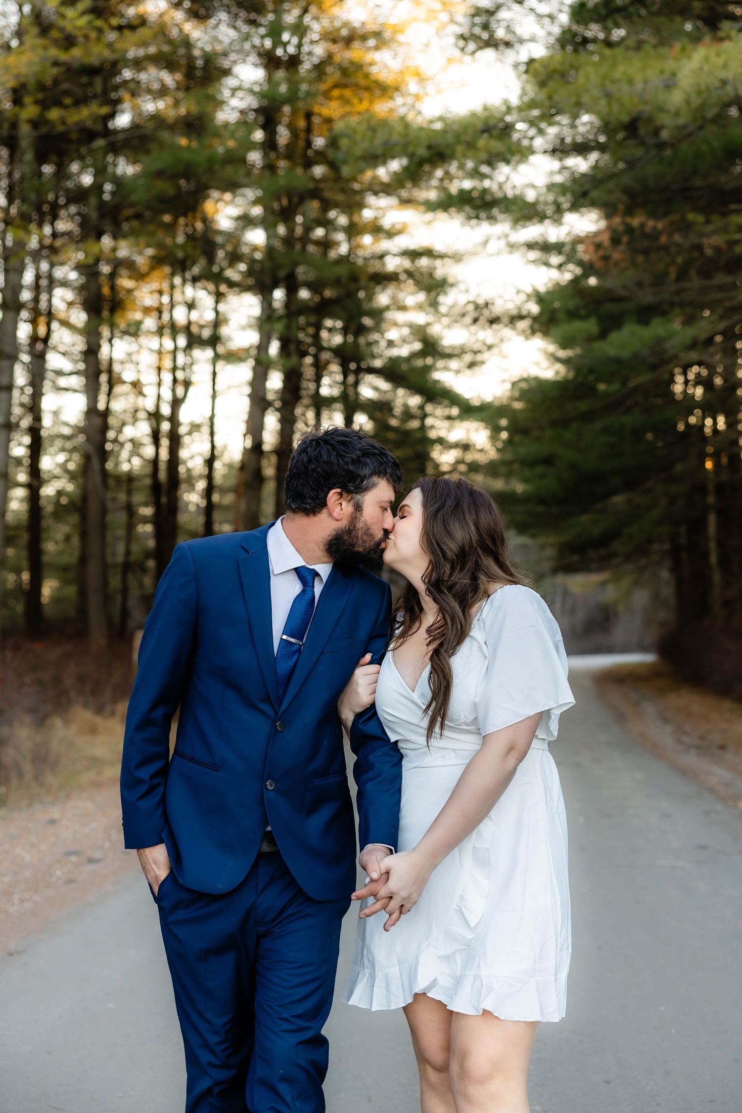 A couple shares an intimate moment on a wooded path, dressed in formal blue suit and white dress.