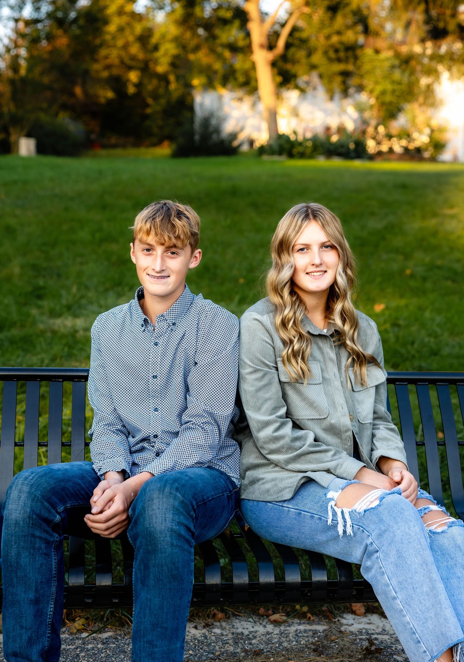 Two siblings sit together on park bench during golden hour photo session.