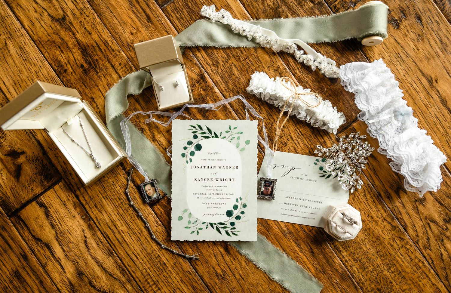 Wedding invitations and accessories arranged on wooden surface with floral design and sage green accents.
