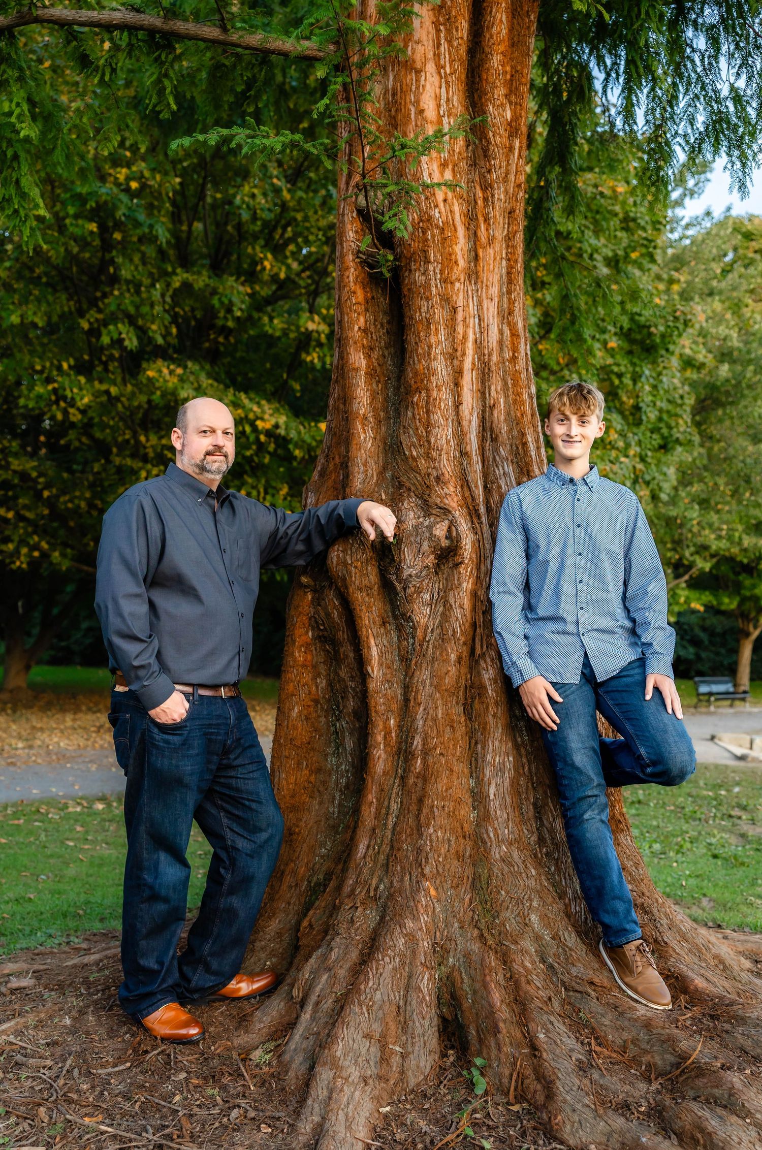 Two pairs of people in complementary outfits pose by large tree trunk in park setting.