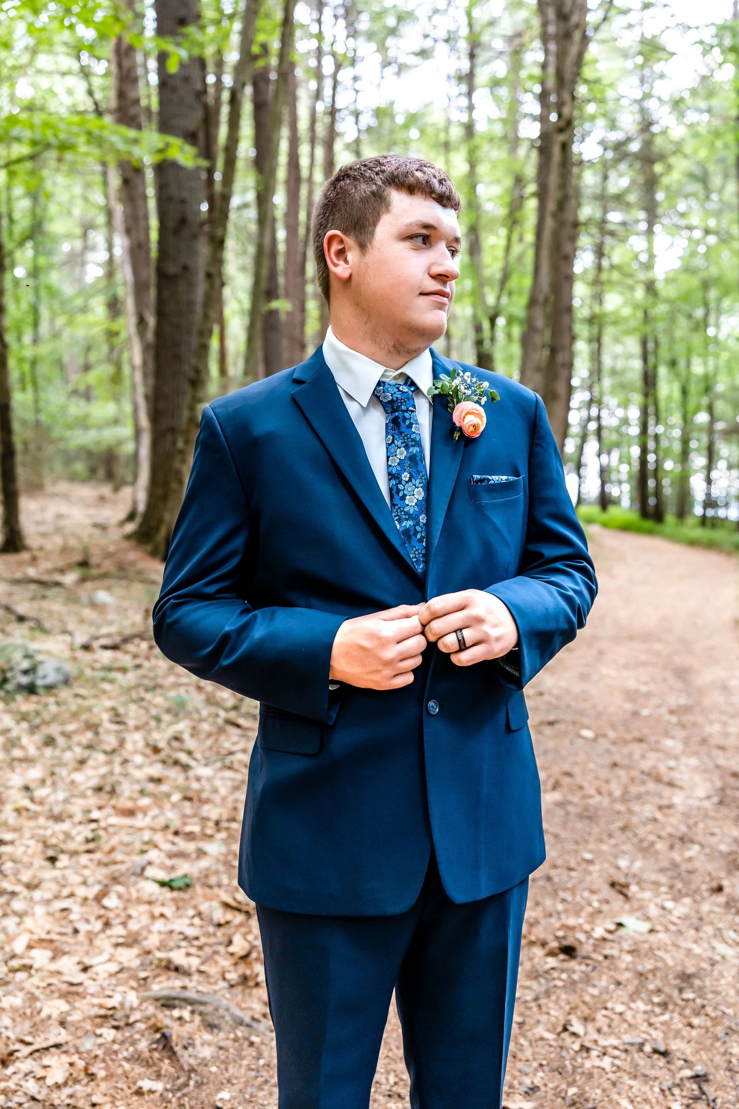 A groom in a navy blue suit and floral tie stands on a forest path.