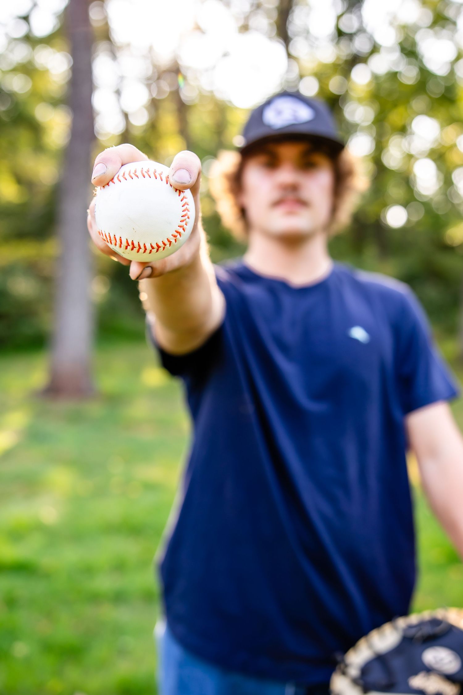 Baseball player in navy blue shirt holding up a baseball in a sunny outdoor setting.