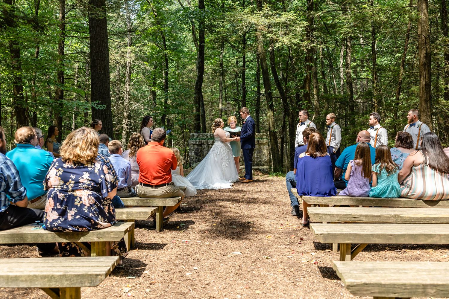 Outdoor forest wedding ceremony with guests seated on wooden benches during the service.
