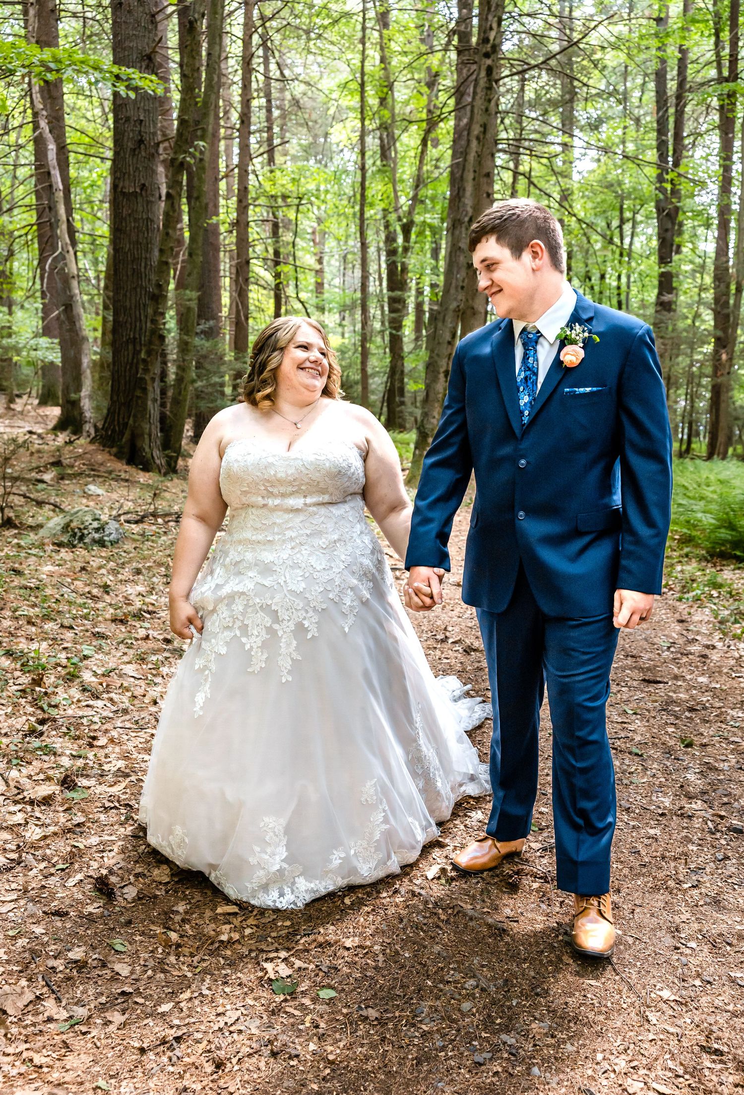 Couple walks hand in hand on forest path in wedding attire.