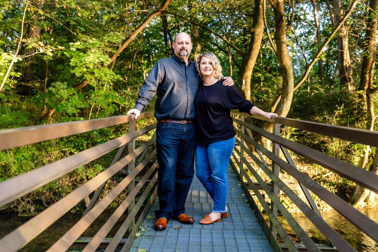 Couple standing together on wooden bridge with forest backdrop.