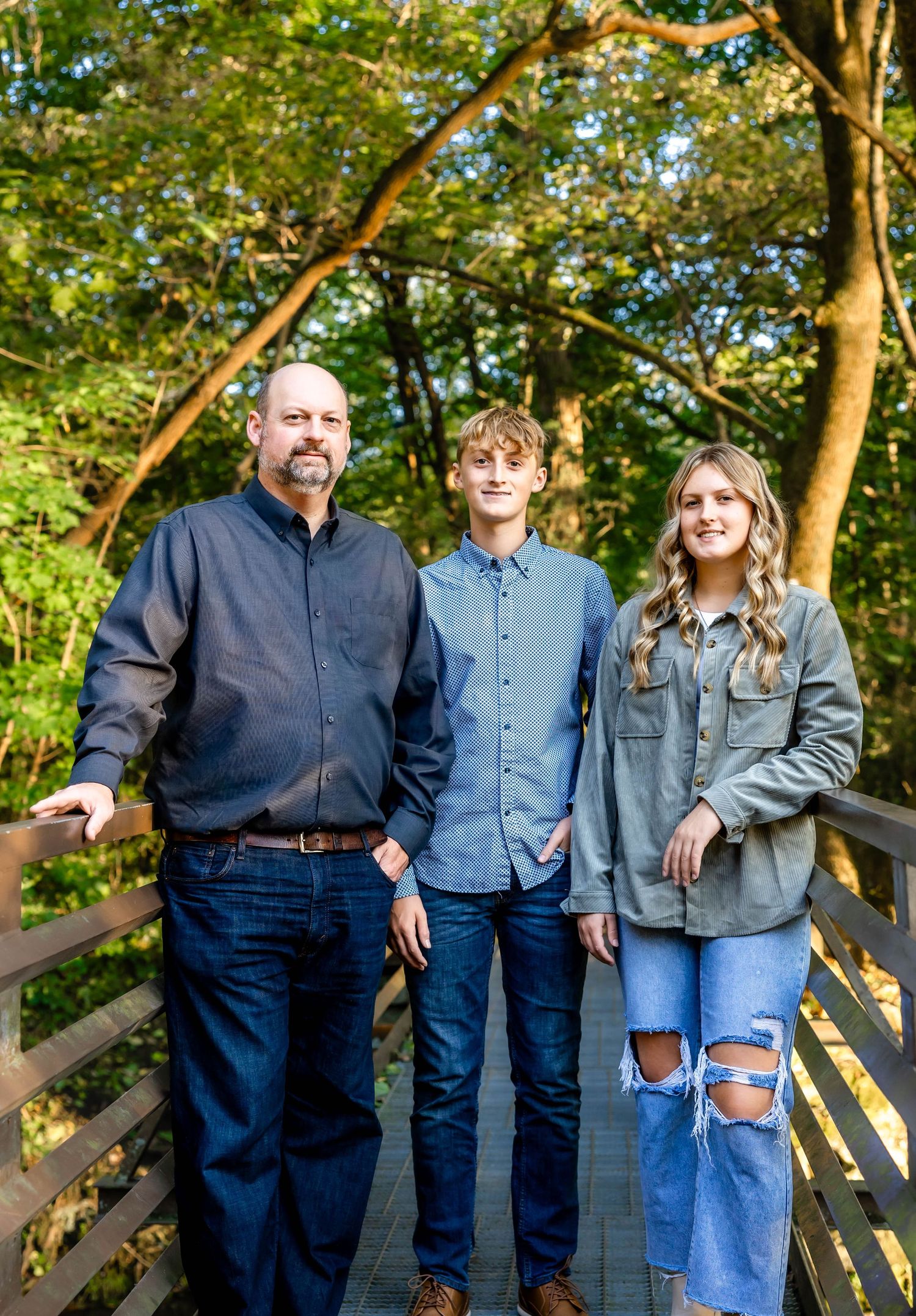 Family of three in coordinated blue outfits poses on wooden boardwalk in forest setting.