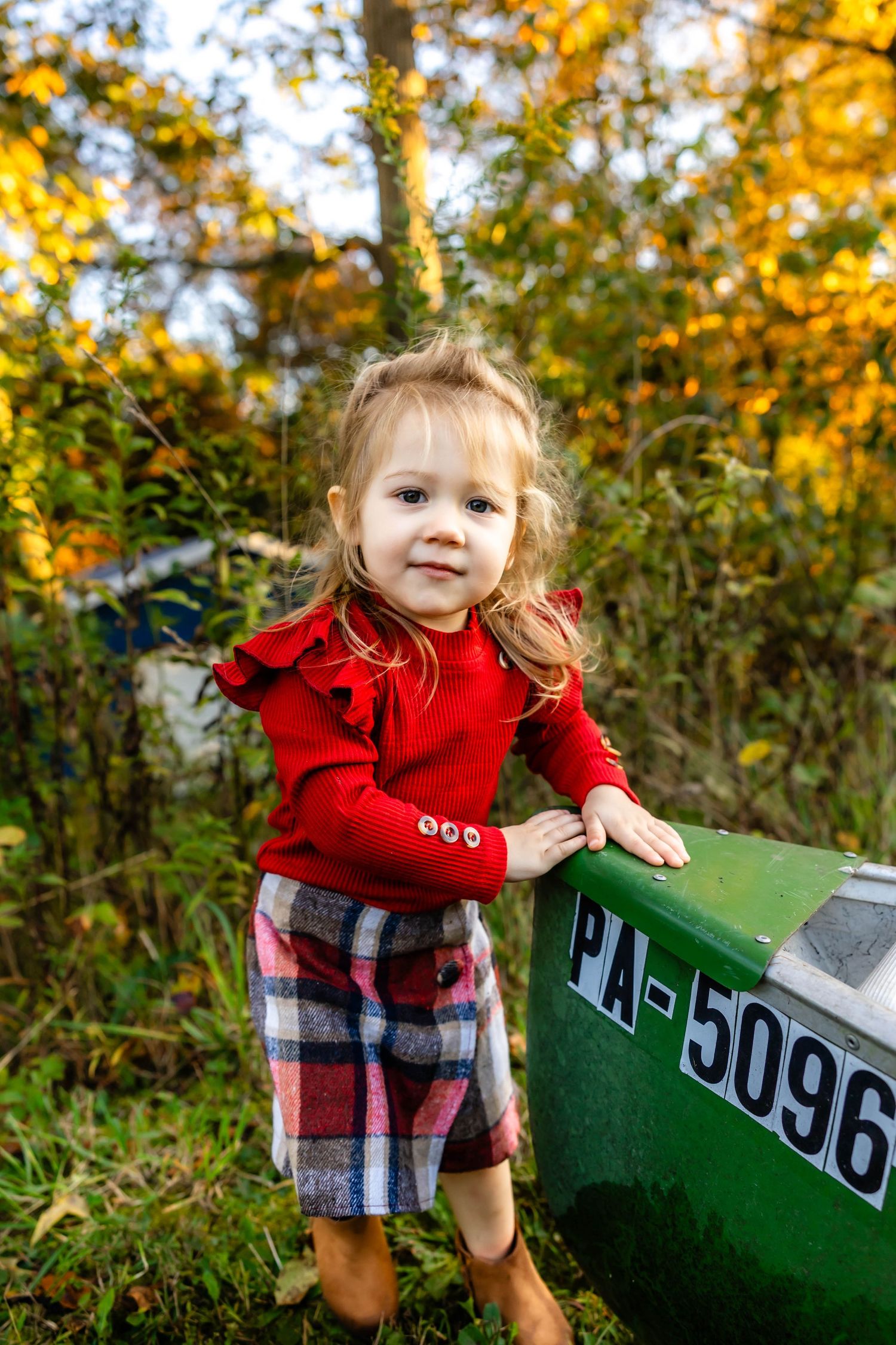 Small child in red sweater and plaid shorts stands next to green farm equipment in autumn setting.