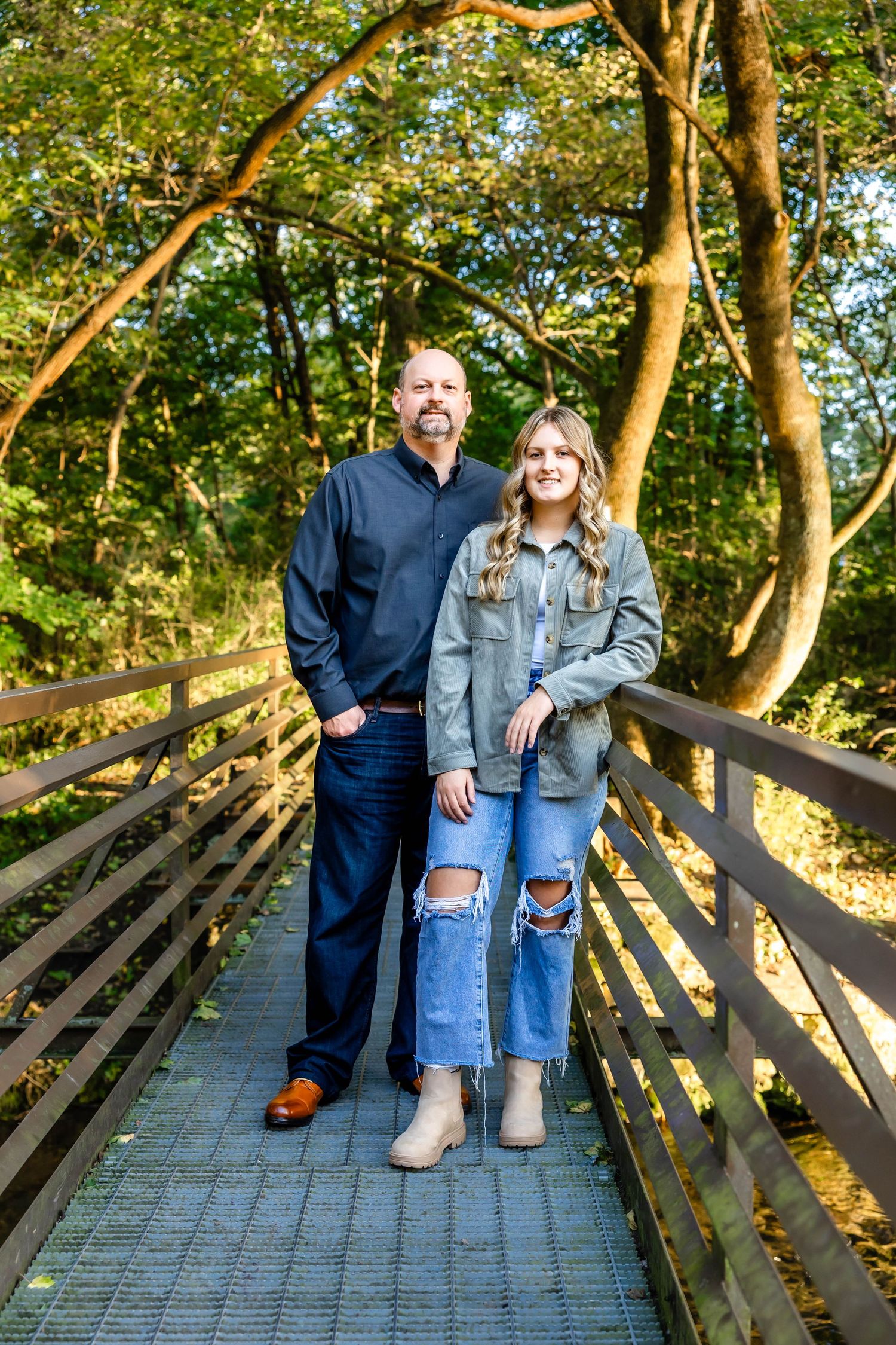 Two people in contrasting casual outfits stand on wooden bridge with sunlit forest backdrop.