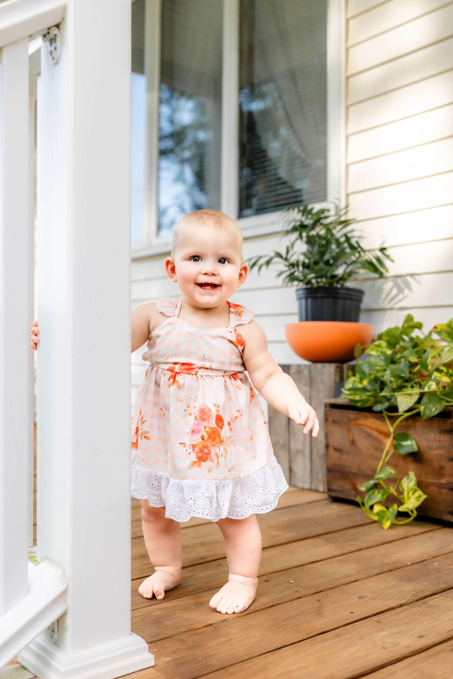 Baby in floral dress standing on wooden porch with planted flowers in background.