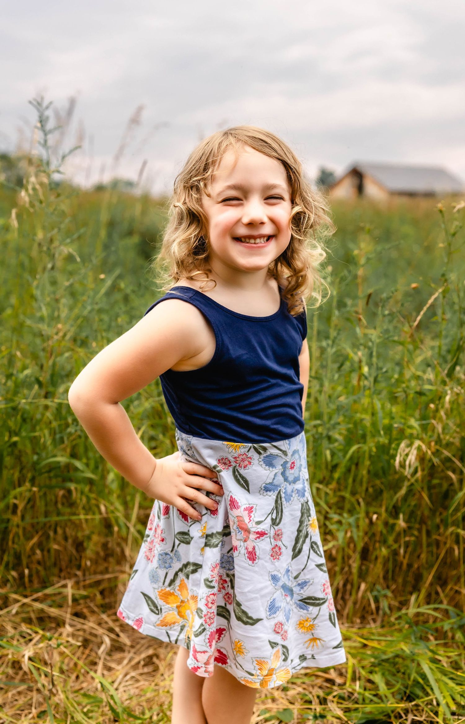 A child in a blue tank top and floral skirt poses in a meadow with tall grasses.
