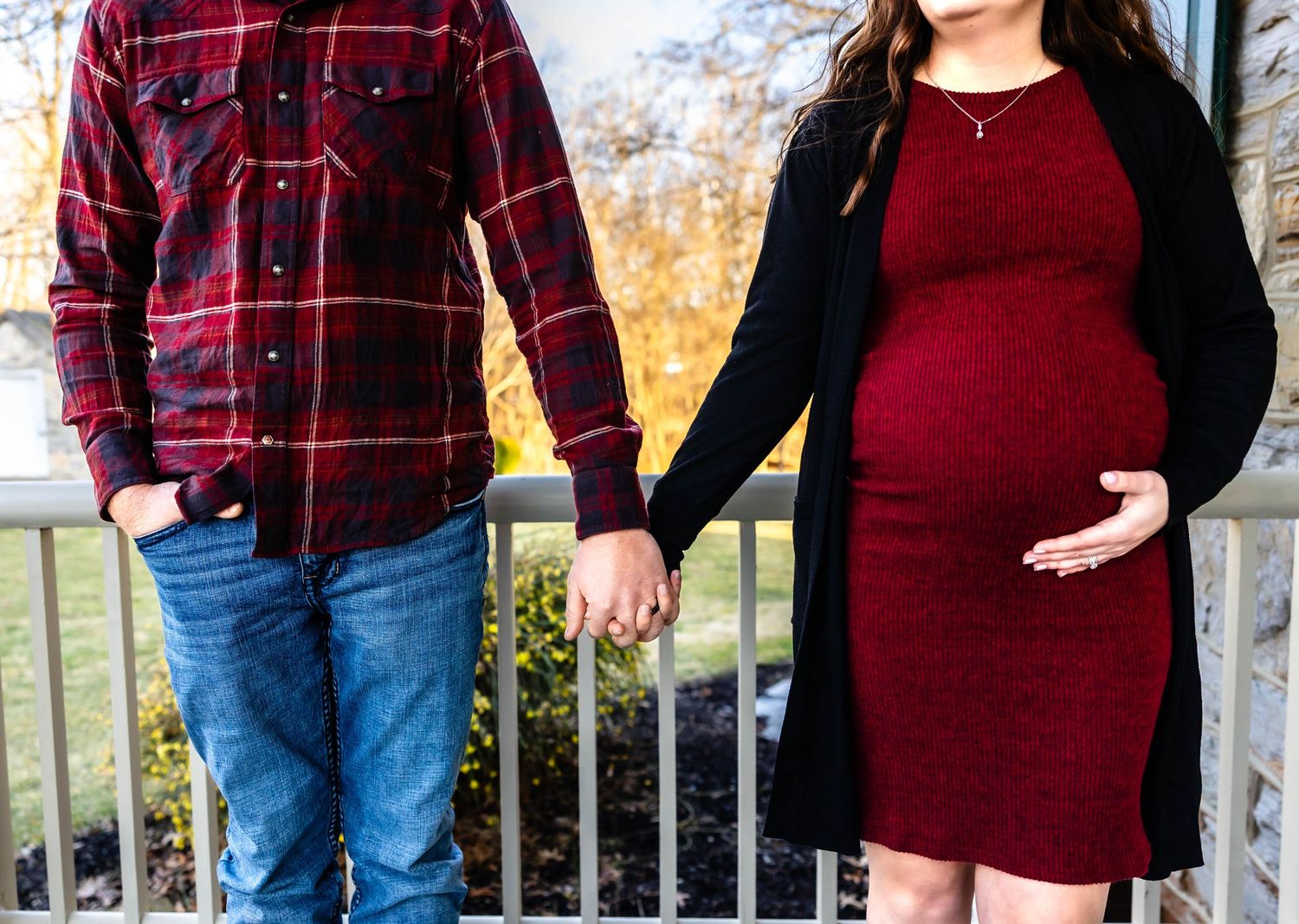 A couple holds hands on a deck wearing coordinating red and black outfits for maternity photos.