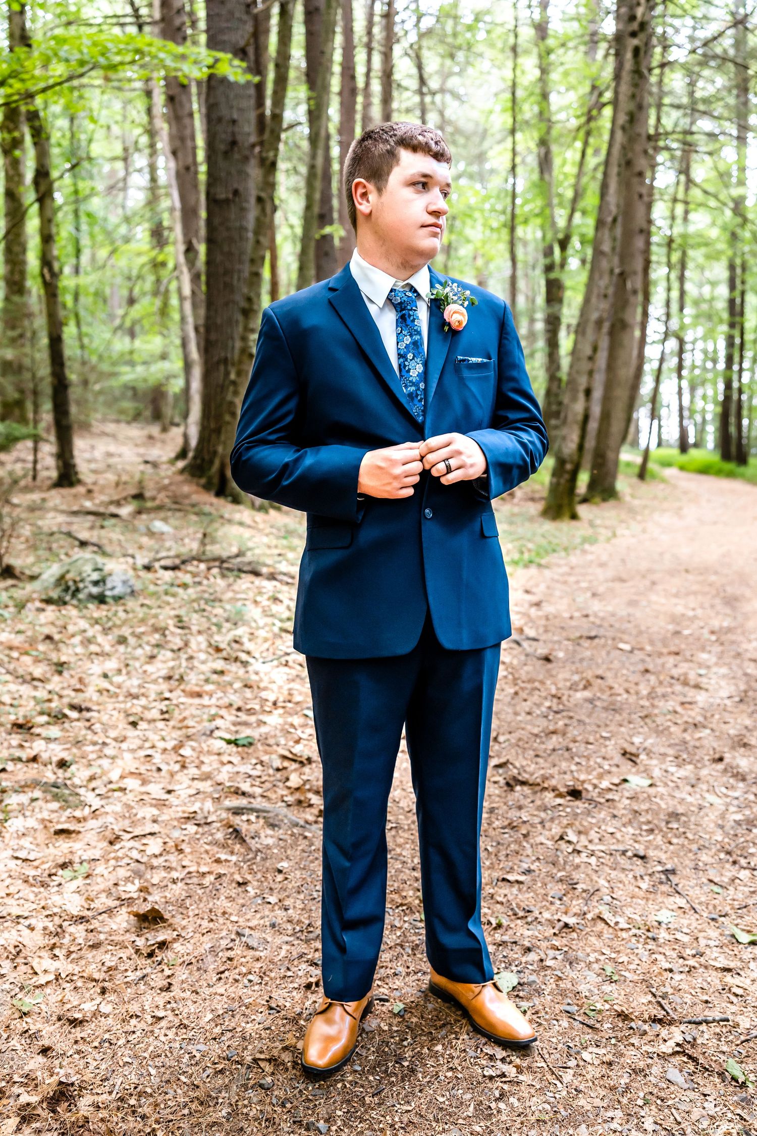 A groom in a navy blue suit and floral tie poses in a wooded forest setting.