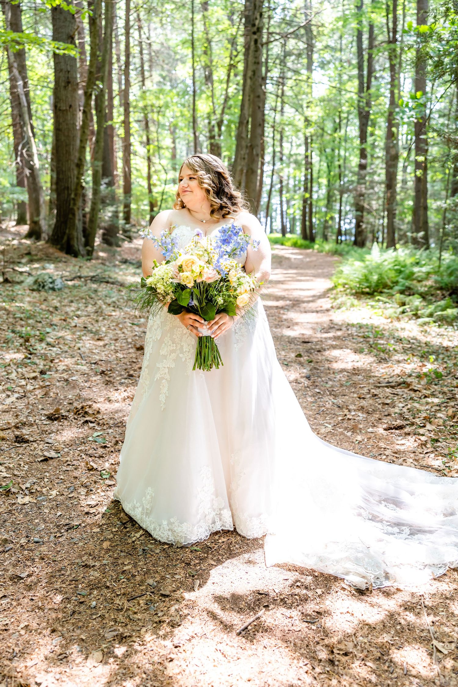 A bride in white lace holds a yellow and white bouquet on a sunlit forest path.