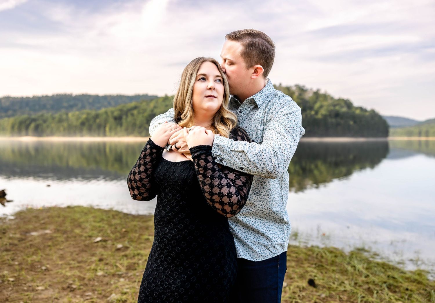 A playful moment between a couple by a lake, with the person in grey hugging from behind.