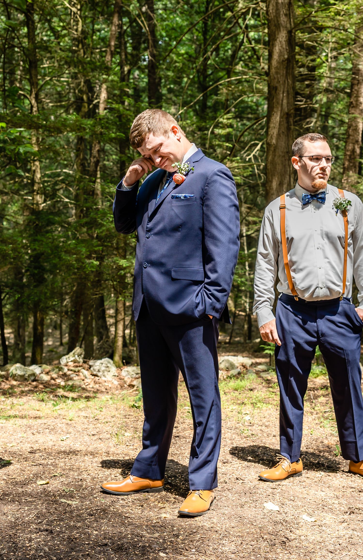 Two wedding attendants in navy suits and suspenders standing in forest clearing.
