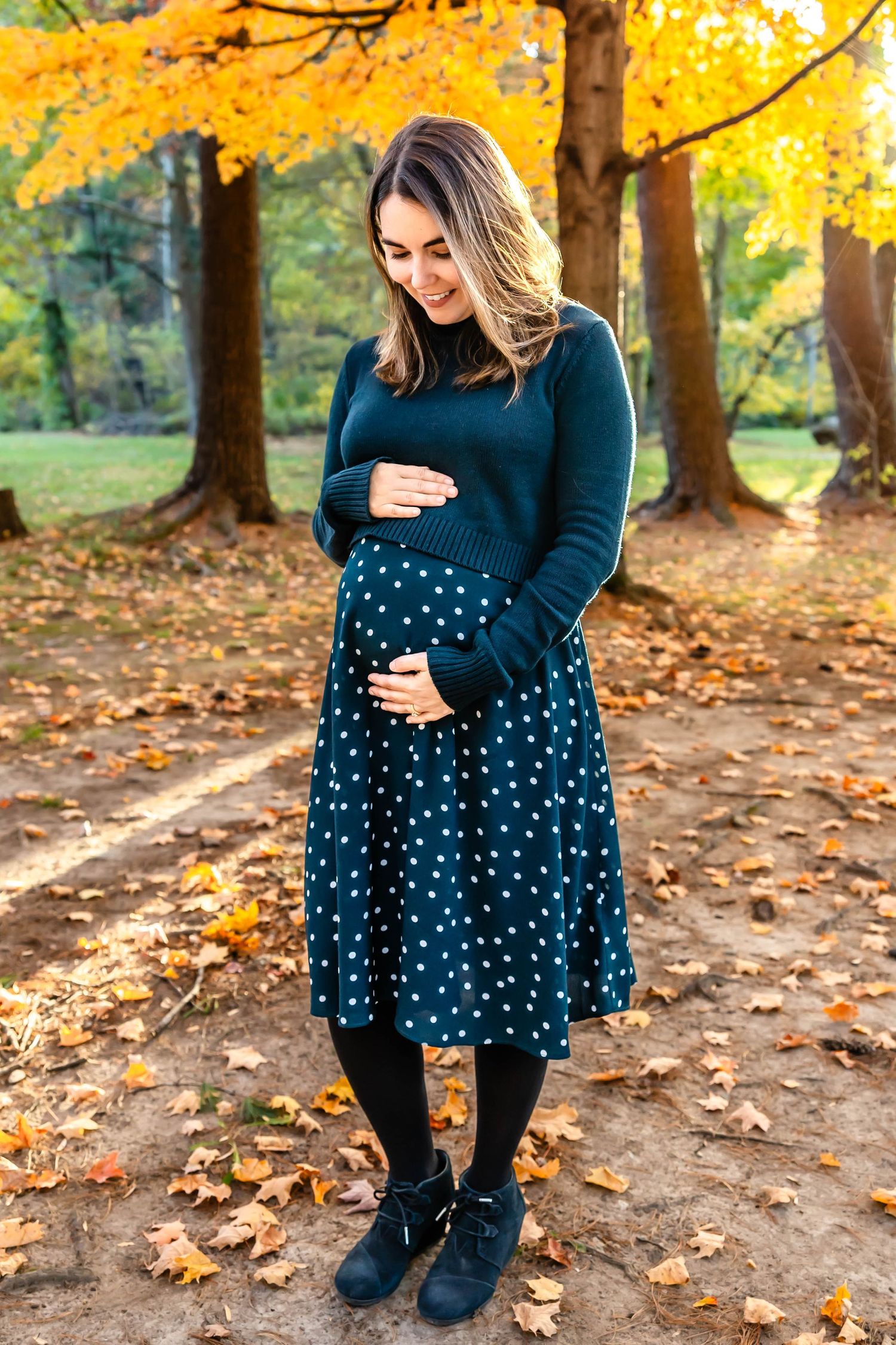 Expectant mother in navy dress smiling while holding her baby bump under autumn trees.