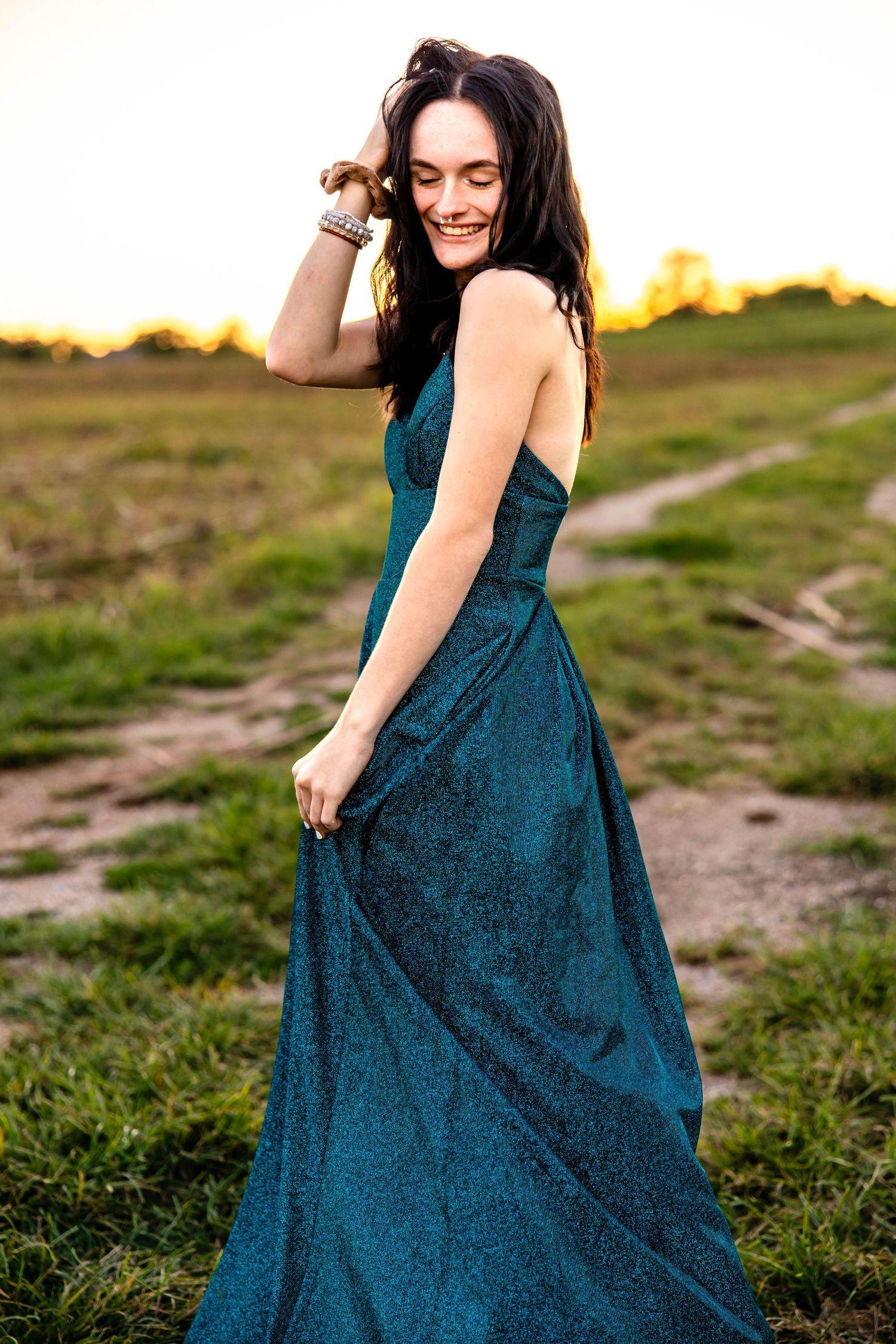 Woman in flowing teal evening gown twirling joyfully at sunset on a dirt path.