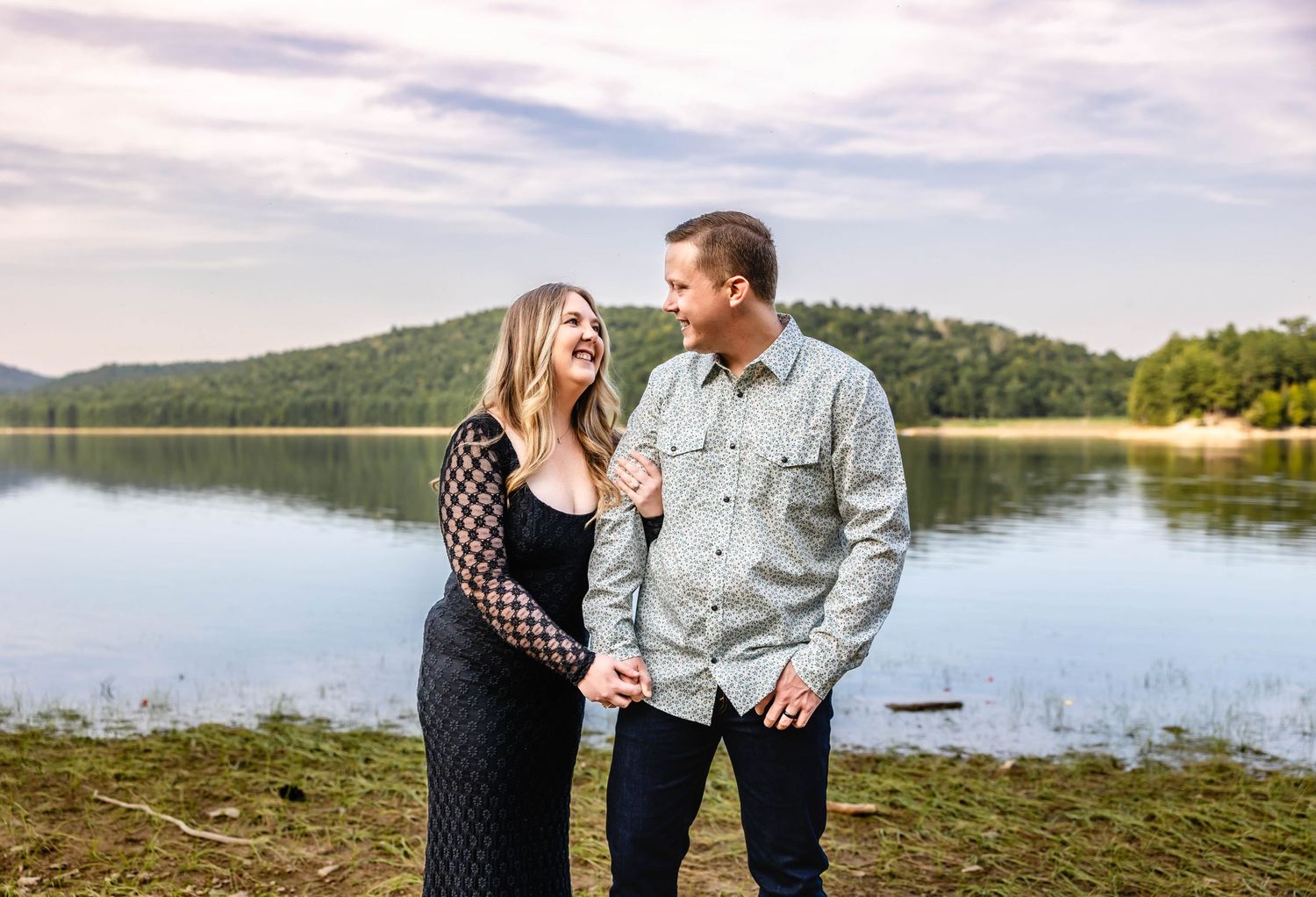 A couple shares a joyful moment by a serene lake with mountains in the background.
