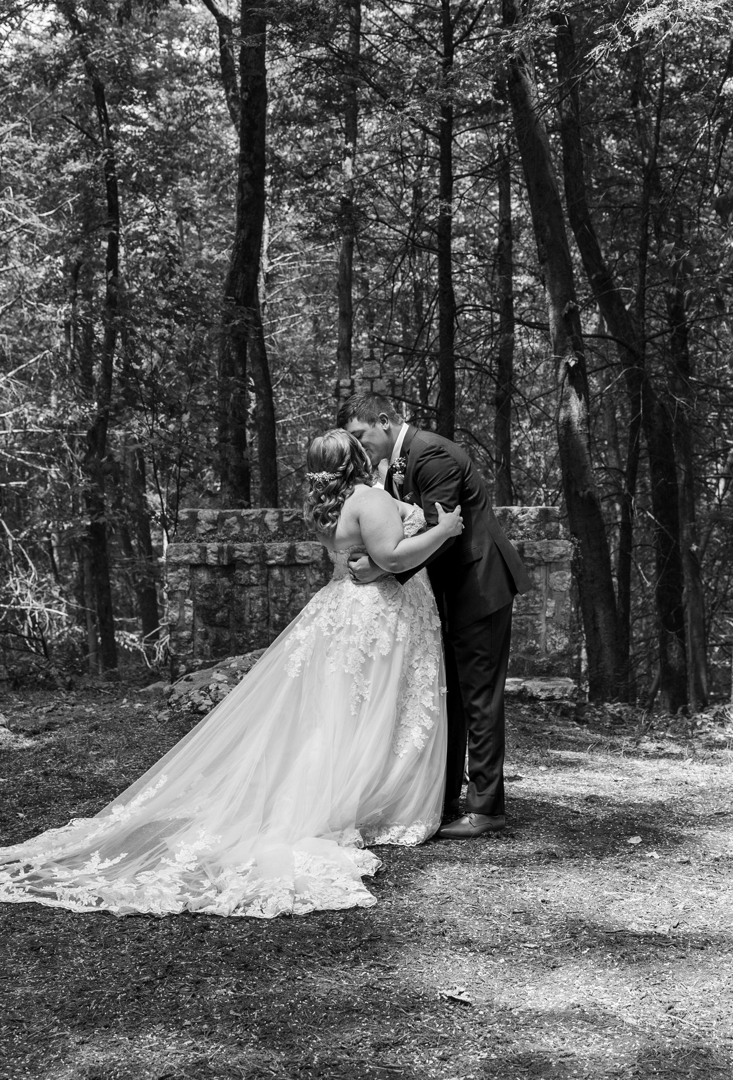 A black and white portrait of a romantic wedding kiss in a wooded setting.