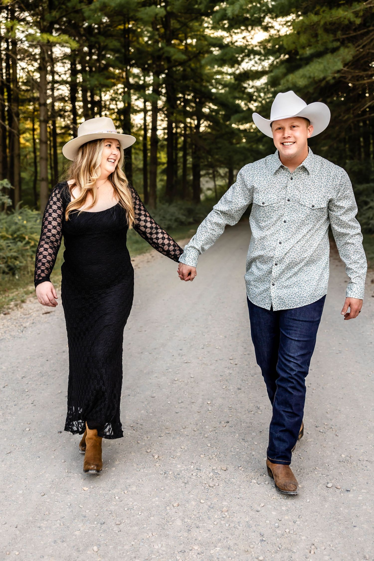 A couple in western wear holding hands while walking down a tree-lined path.
