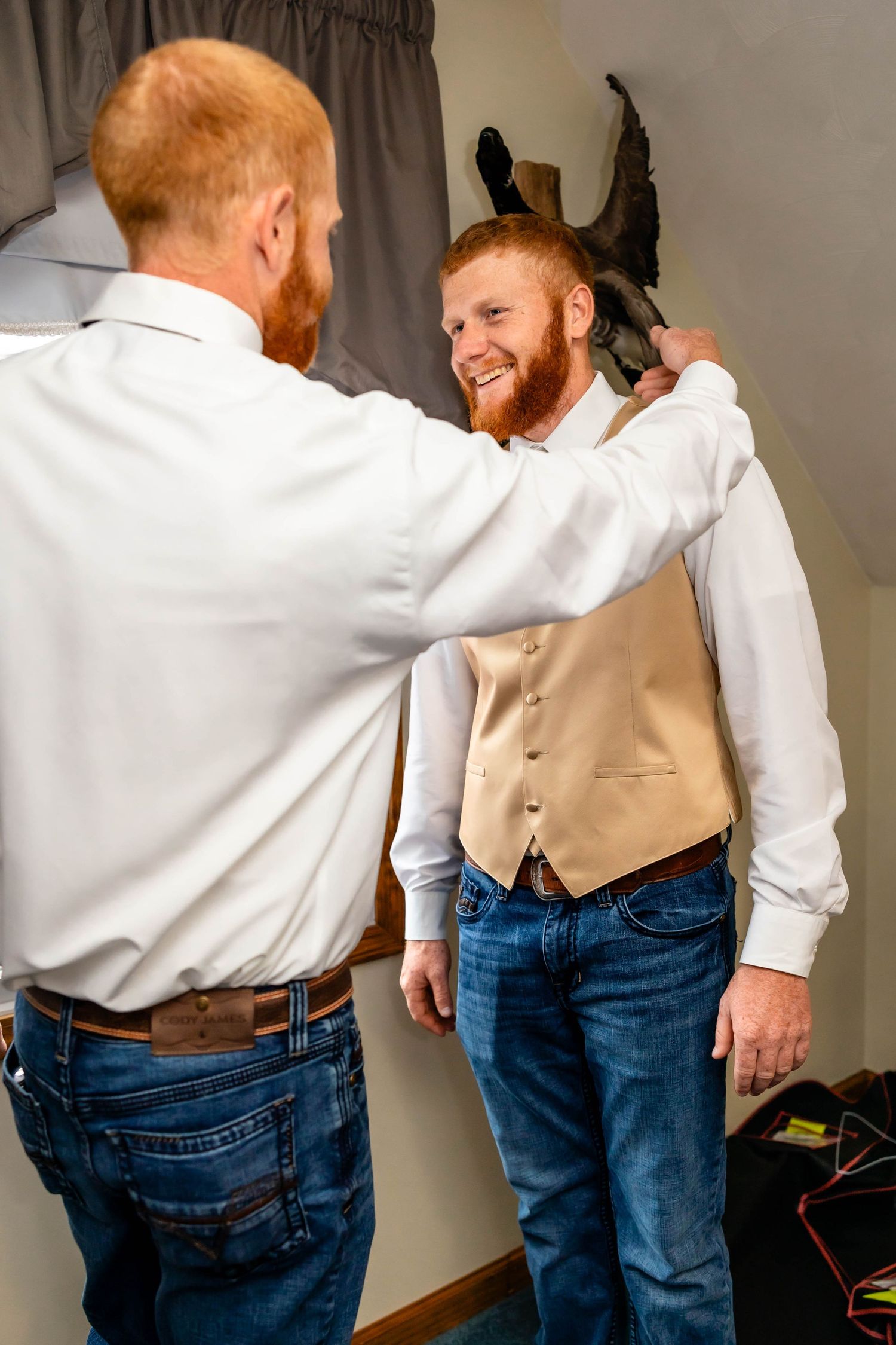 Two people helping with formal attire preparations in a bedroom setting.