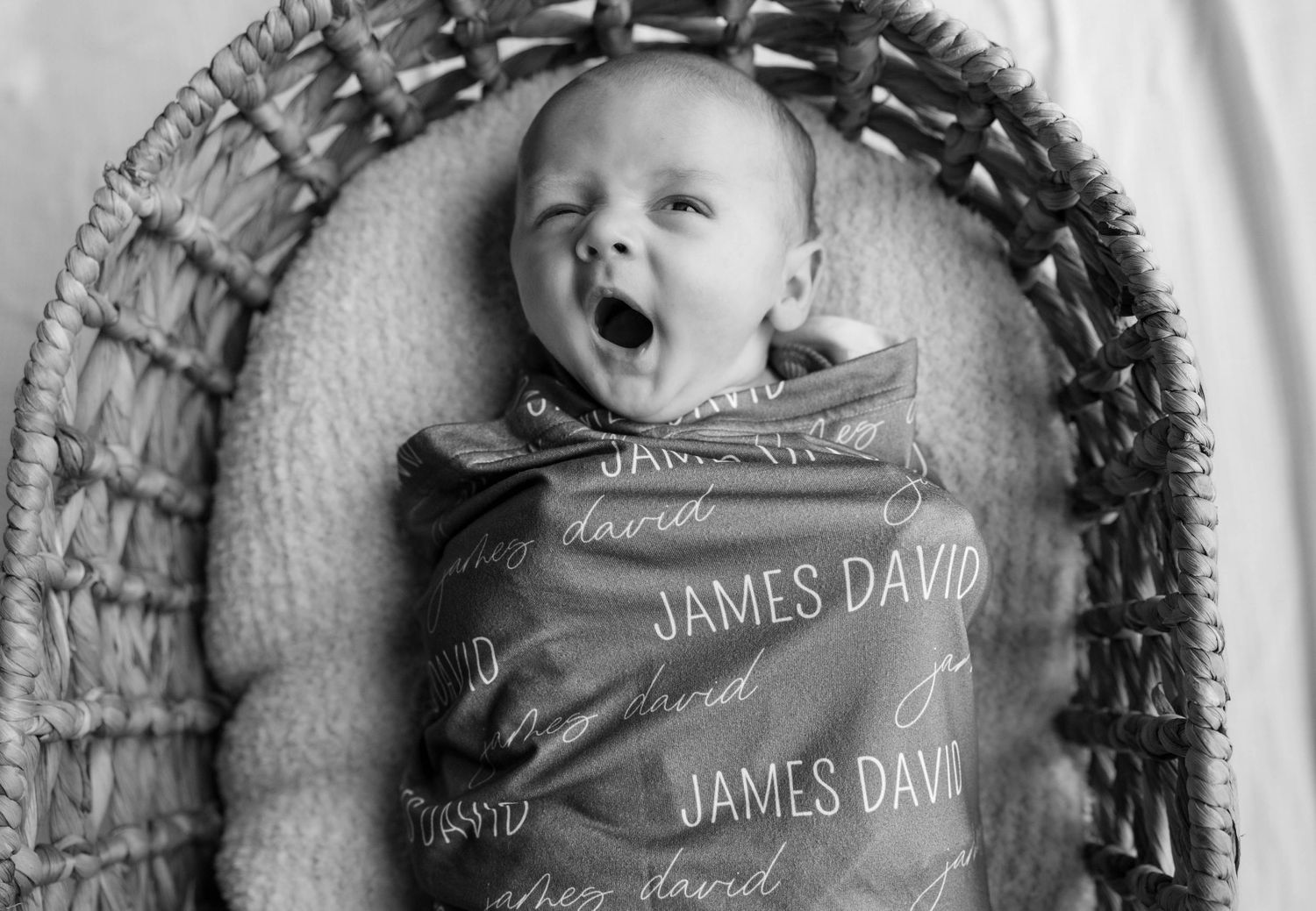 A baby yawns while nestled in a woven basket wrapped in a custom name blanket.