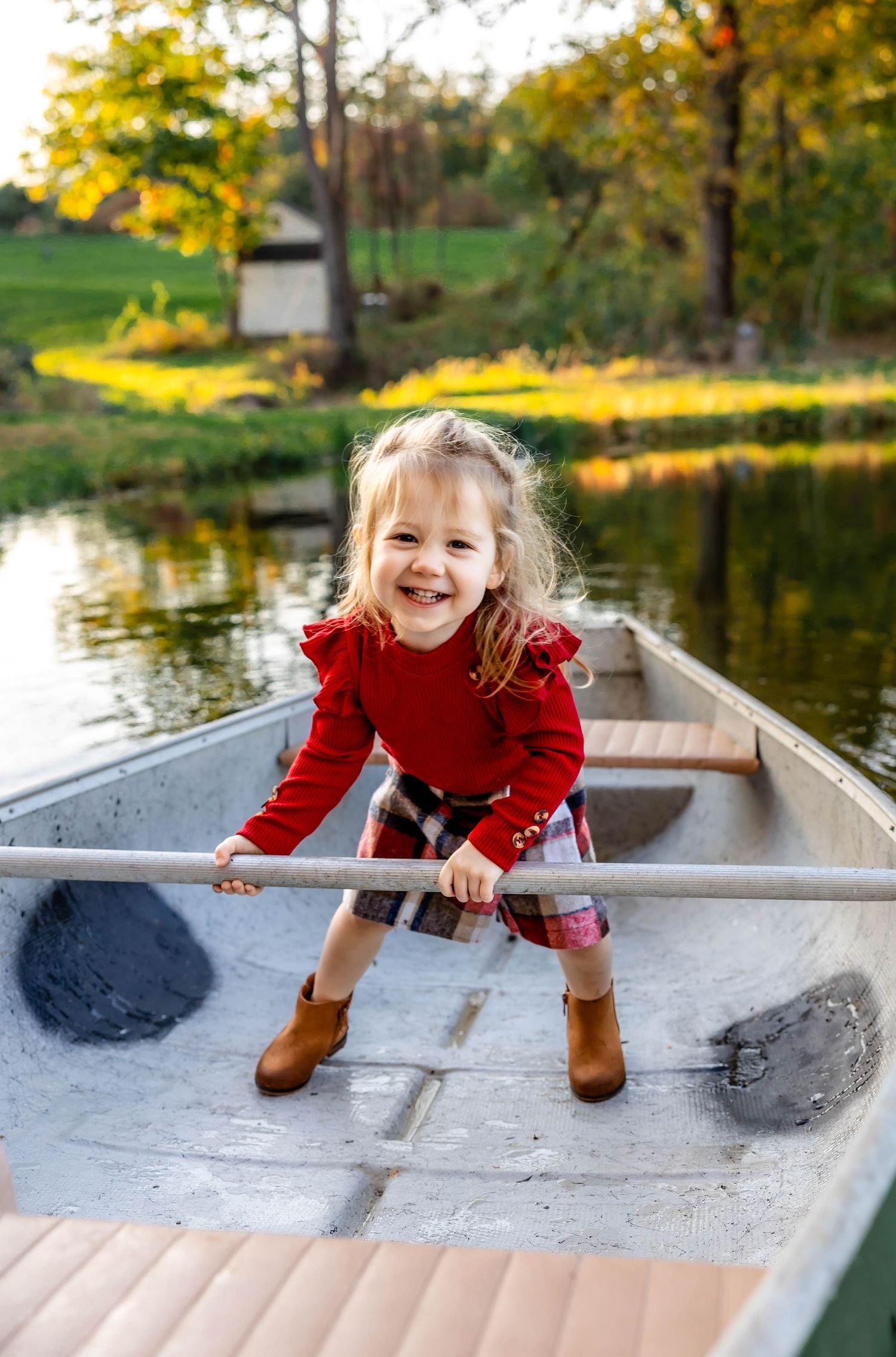 Child in red sweater and plaid skirt holds paddle while sitting in canoe during fall.
