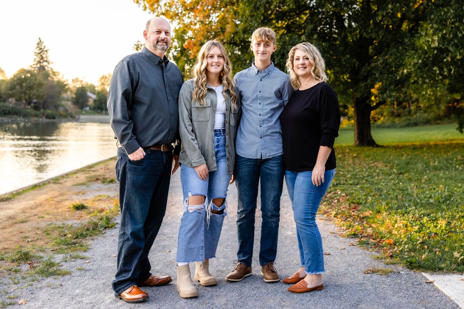 A family stands together on a path near a lake during autumn with trees in the background.