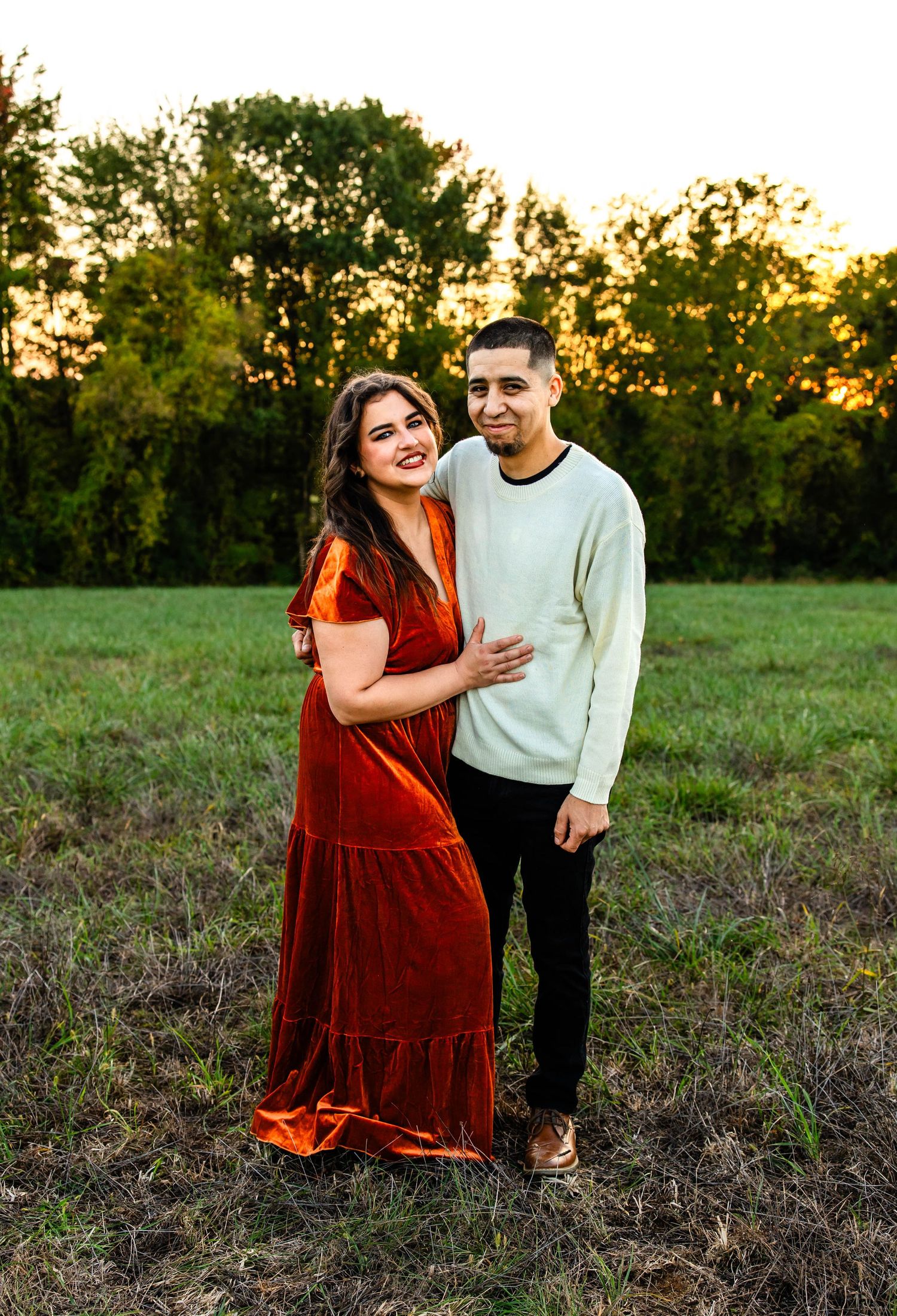 Couple in rustic field at golden hour, wearing white sweater and red velvet dress.