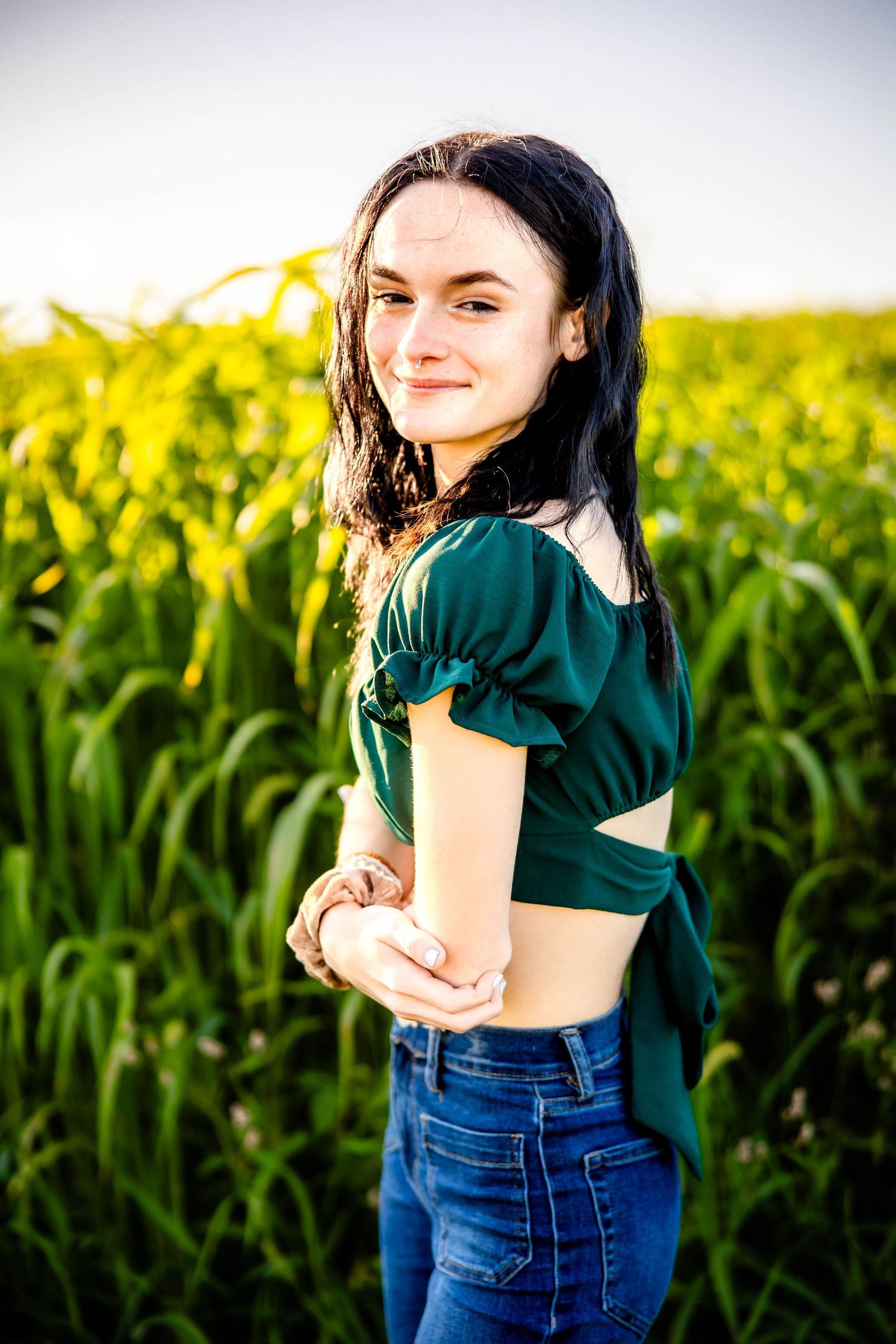 A person in emerald green top and jeans poses against a golden cornfield background.