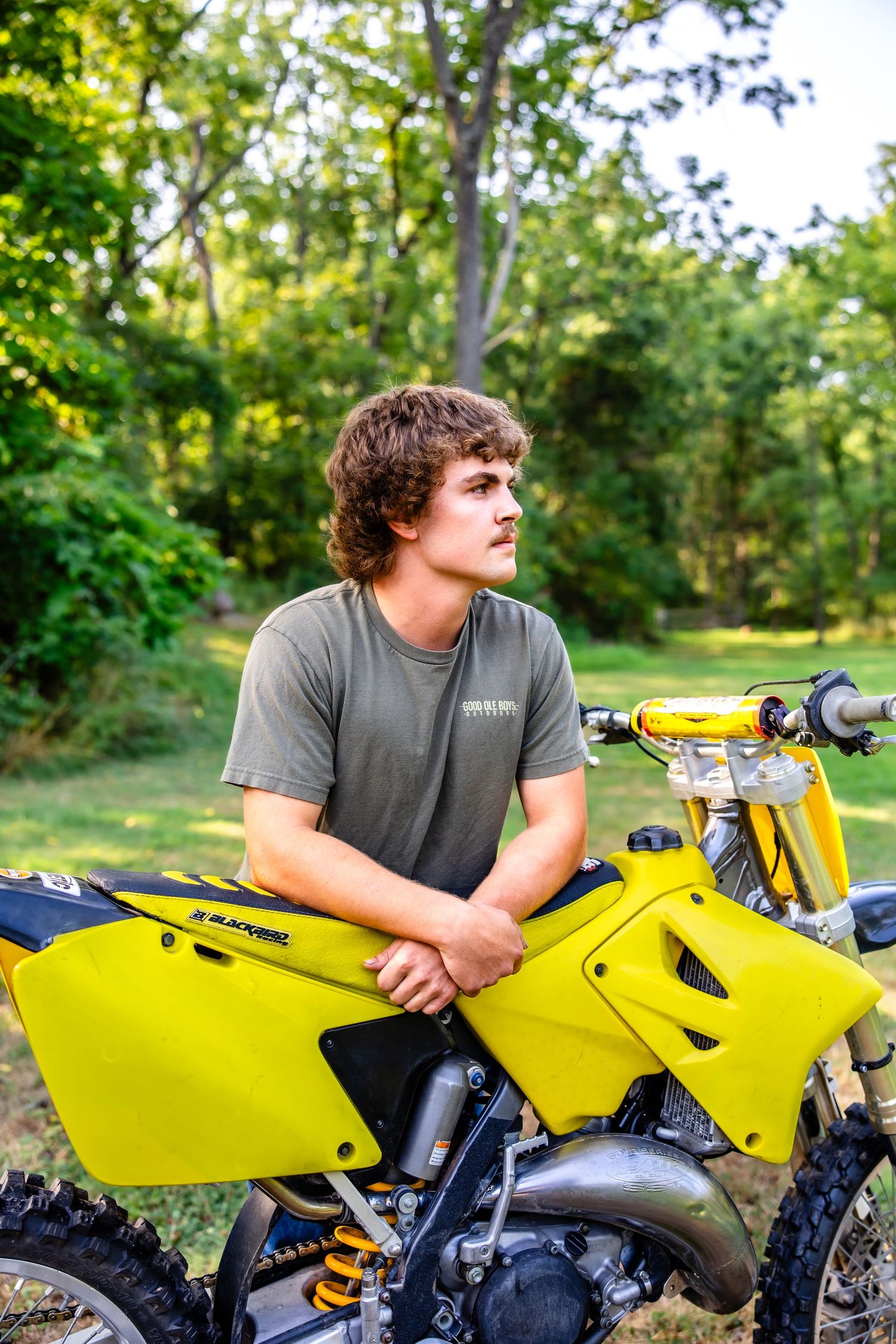 Person in gray t-shirt leans against bright yellow dirt bike in forest setting.