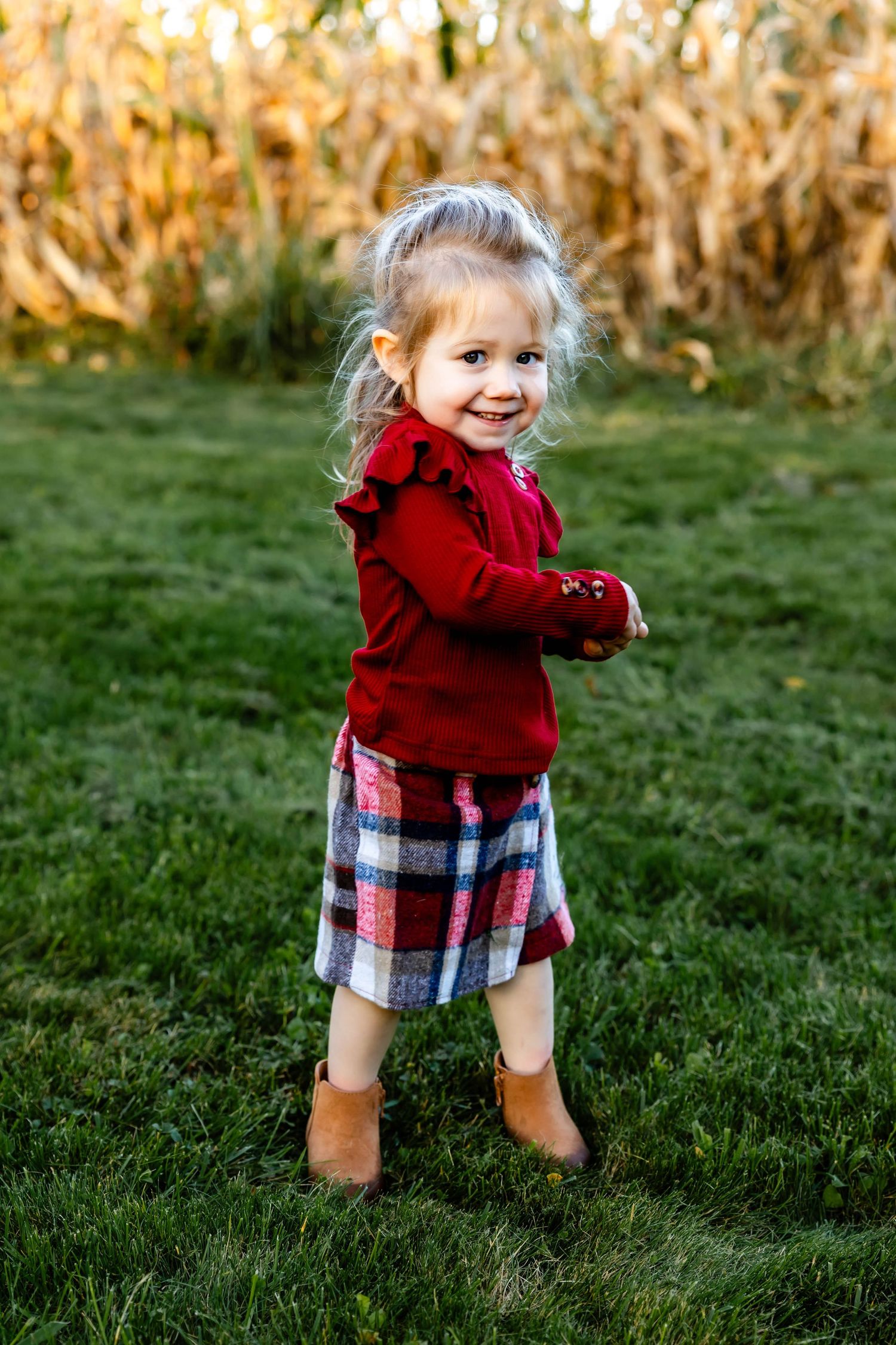 Smiling child in red ruffled sweater and plaid skirt stands on green grass near corn field.