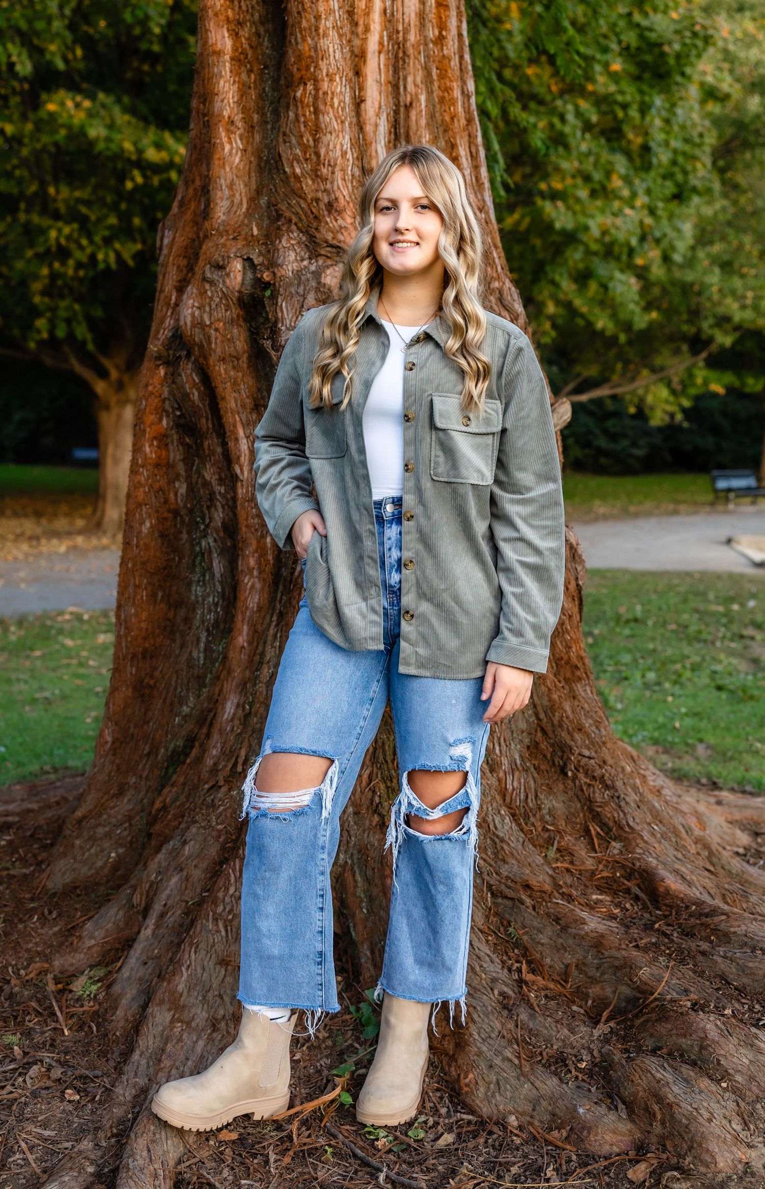 Person wearing olive utility jacket and distressed jeans poses by massive tree trunk in park setting.