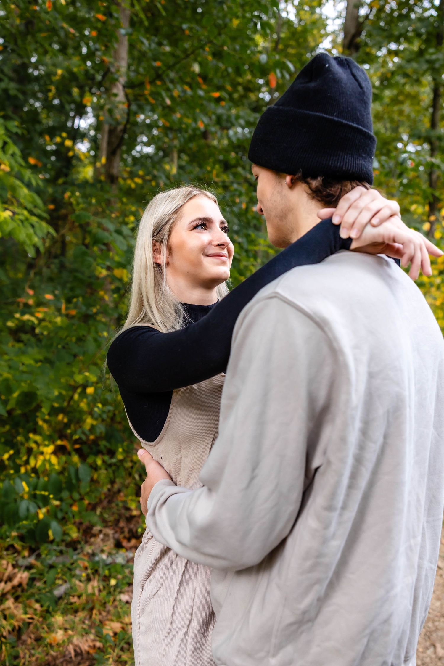 A romantic couple embraces while standing on a forest path with fall colors in the background.