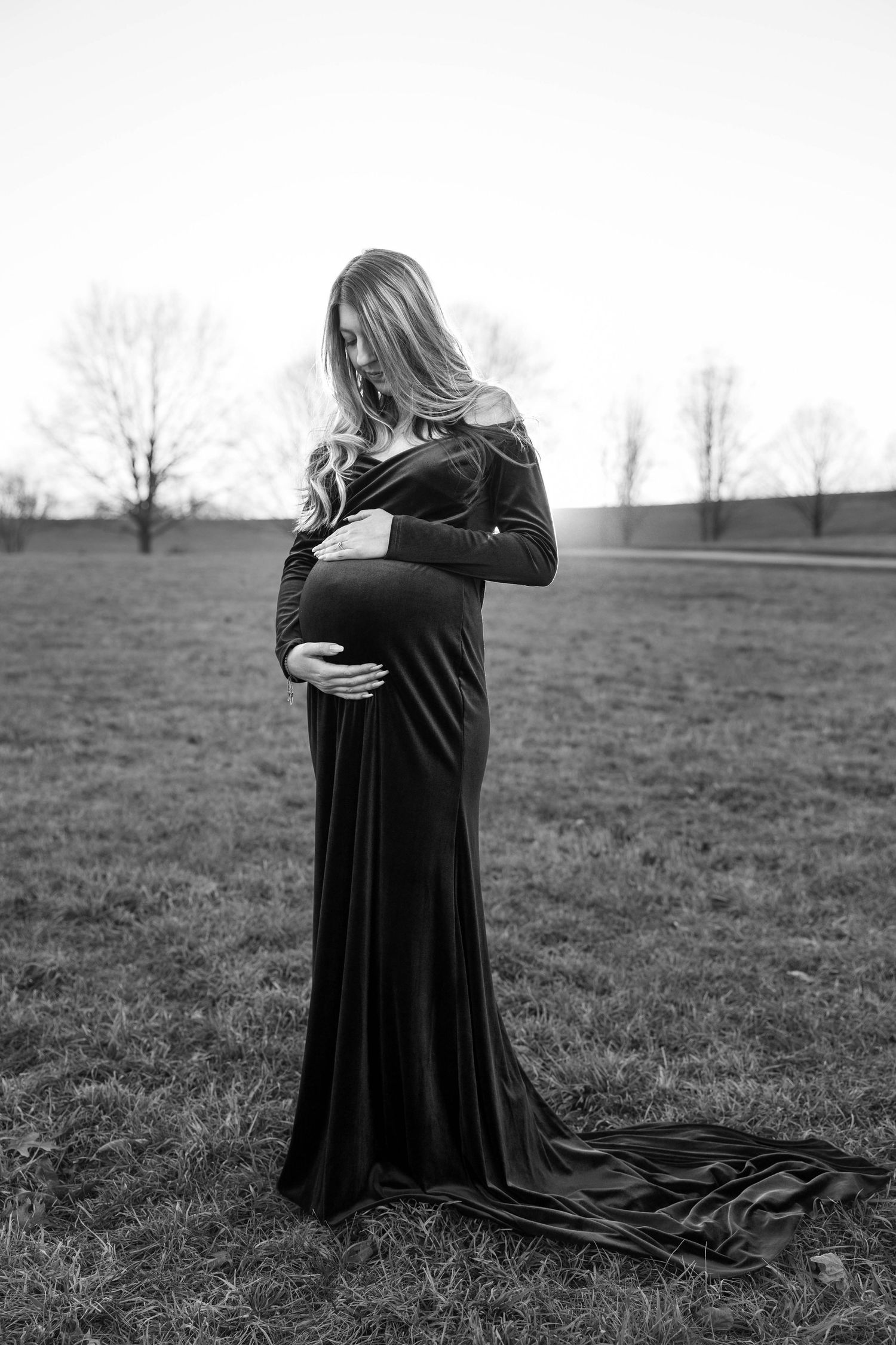 Dramatic black and white maternity photo featuring a long flowing dress against a nature backdrop.