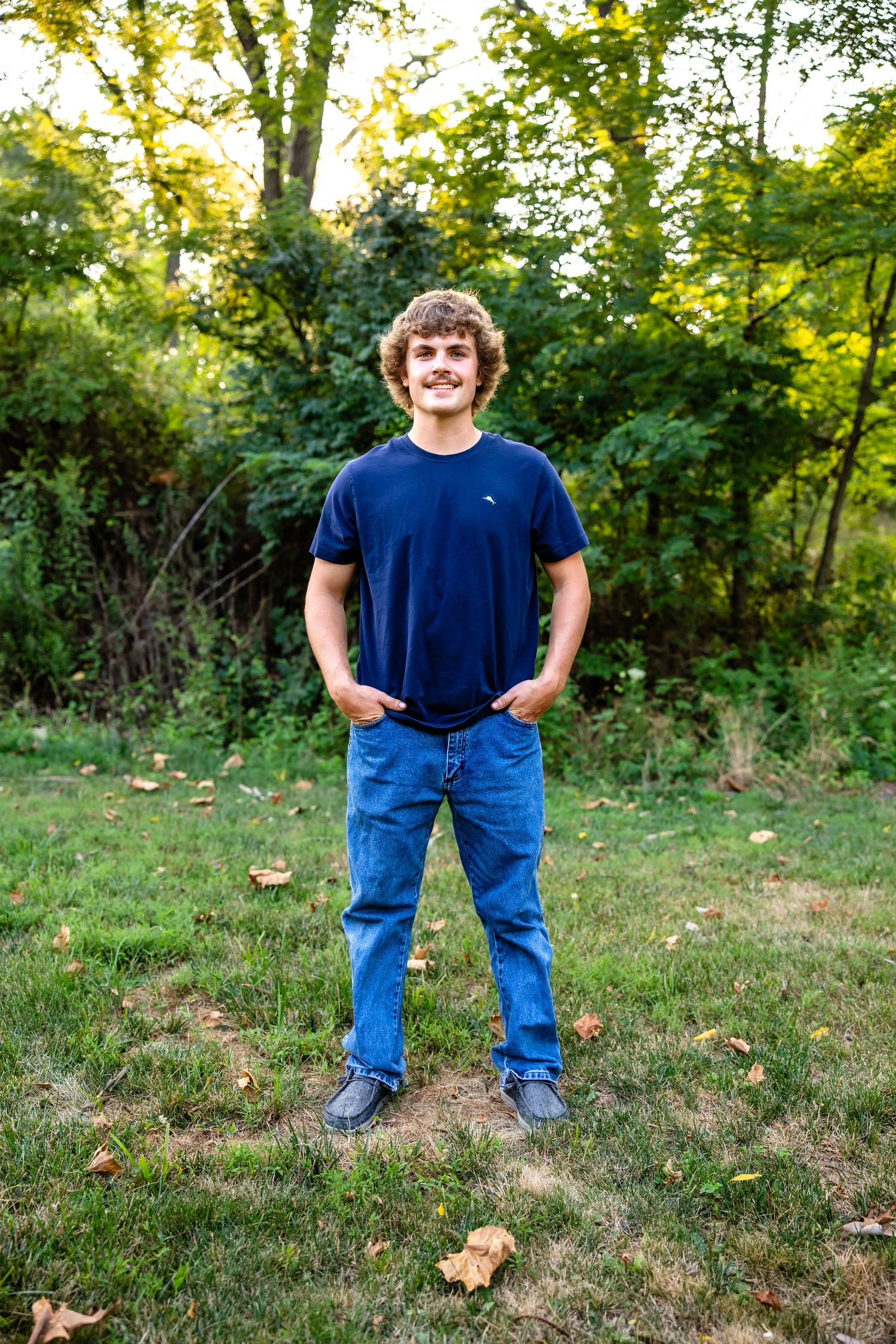 Standing outdoors in navy t-shirt and jeans with autumn trees in background.