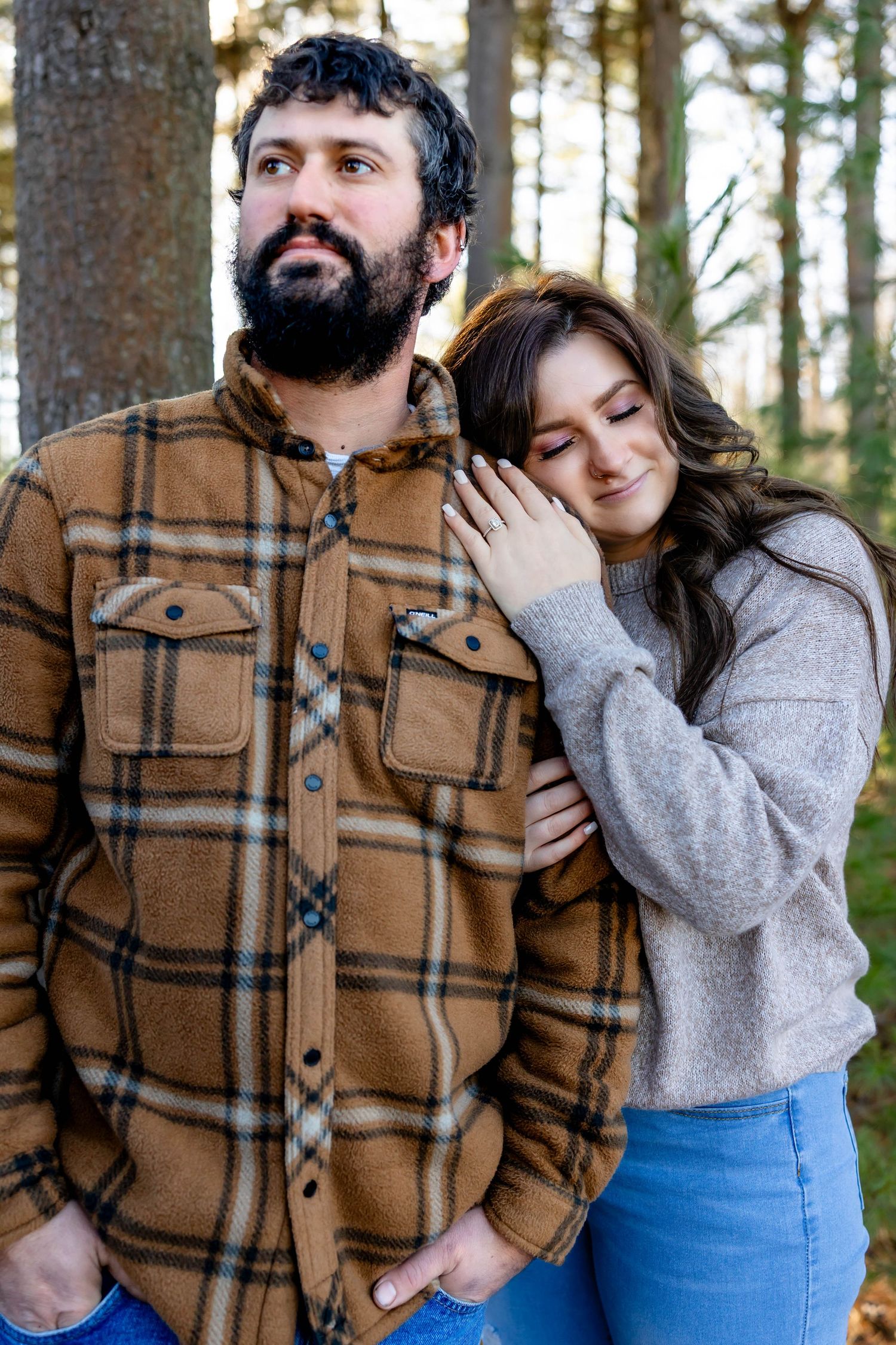 A couple poses together outdoors, with one wearing a plaid flannel and the other in a gray sweater.
