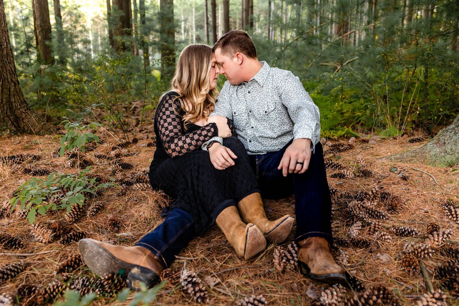 A couple sits together on fallen leaves in a wooded area during an autumn day.