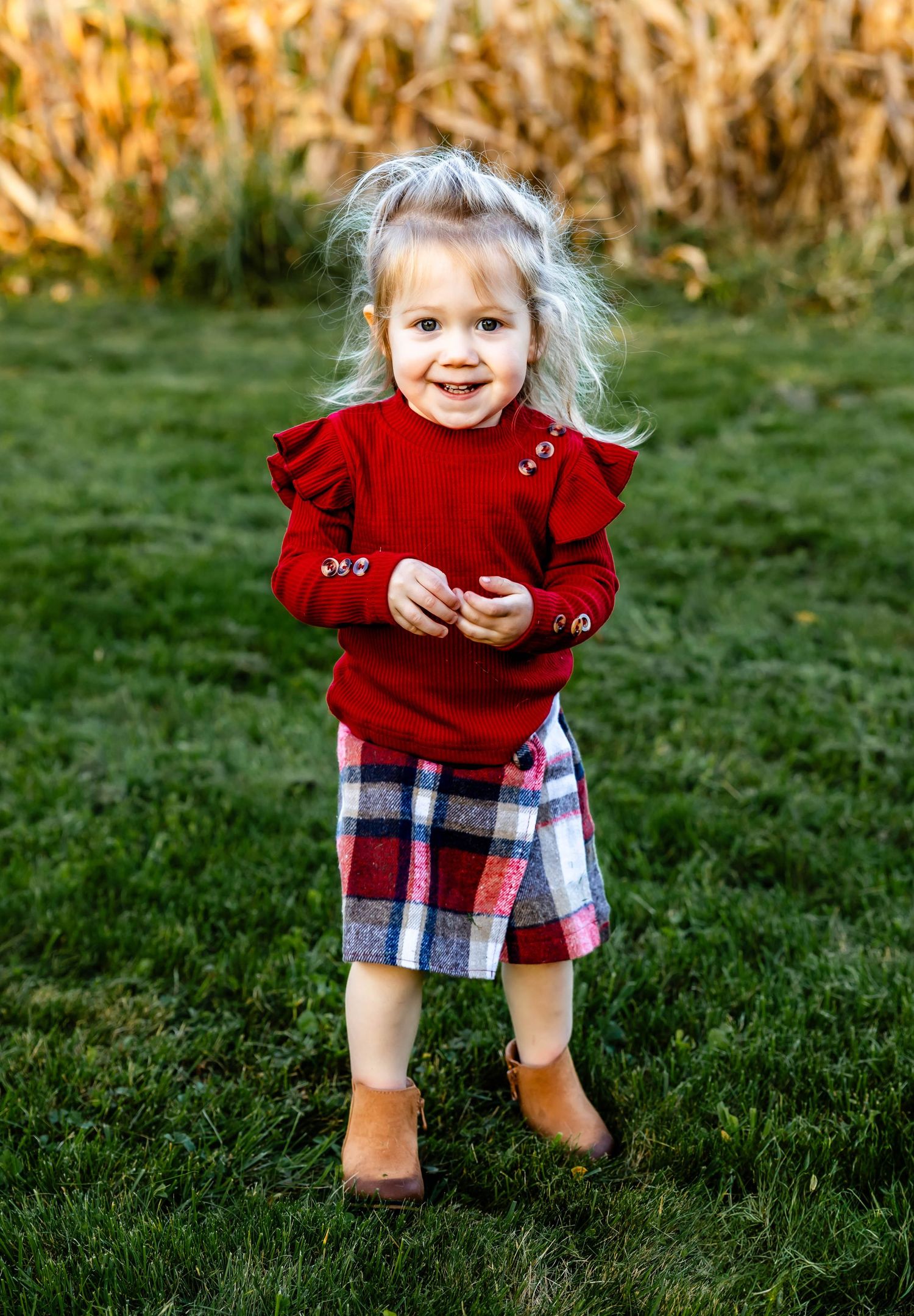 Toddler wearing red sweater and plaid skirt plays in grassy field with autumn corn stalks in background.