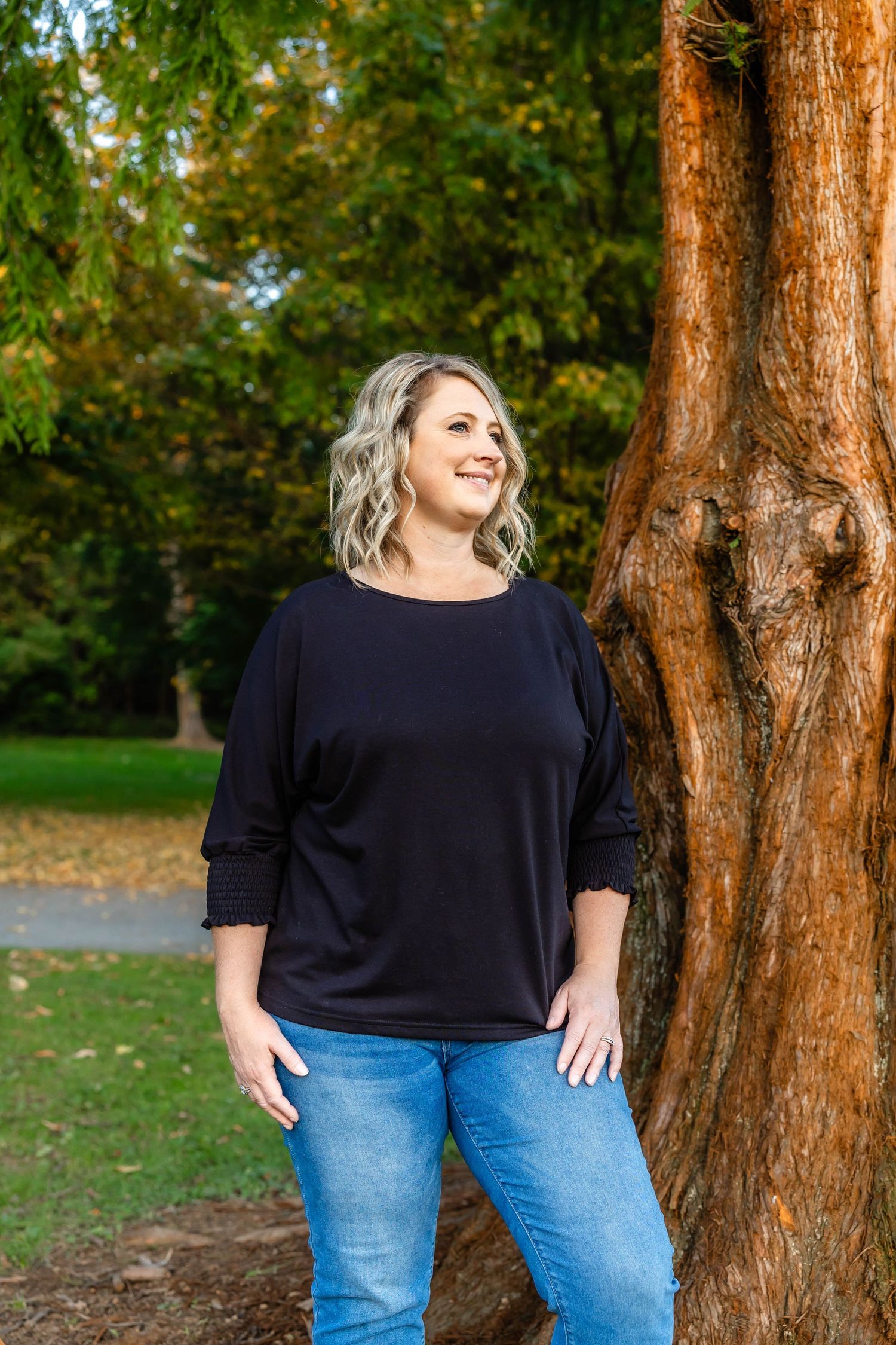 Casual outdoor portrait with person in dark top and jeans standing next to a large redwood tree.