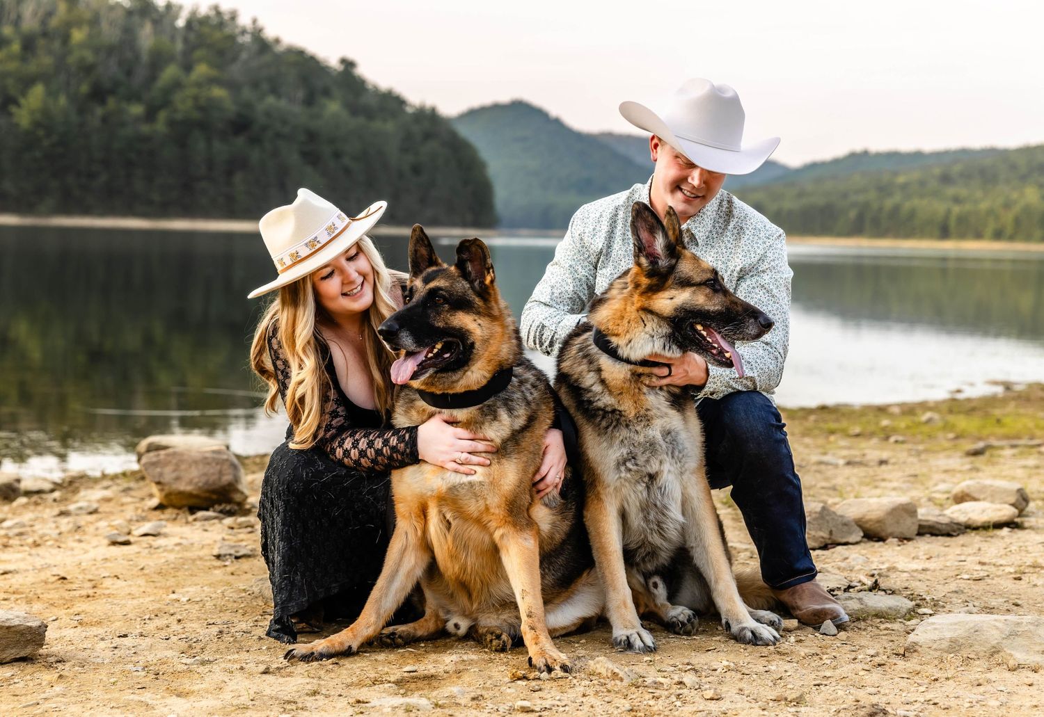 A couple in western wear sits with their German Shepherd dogs by a scenic lake.