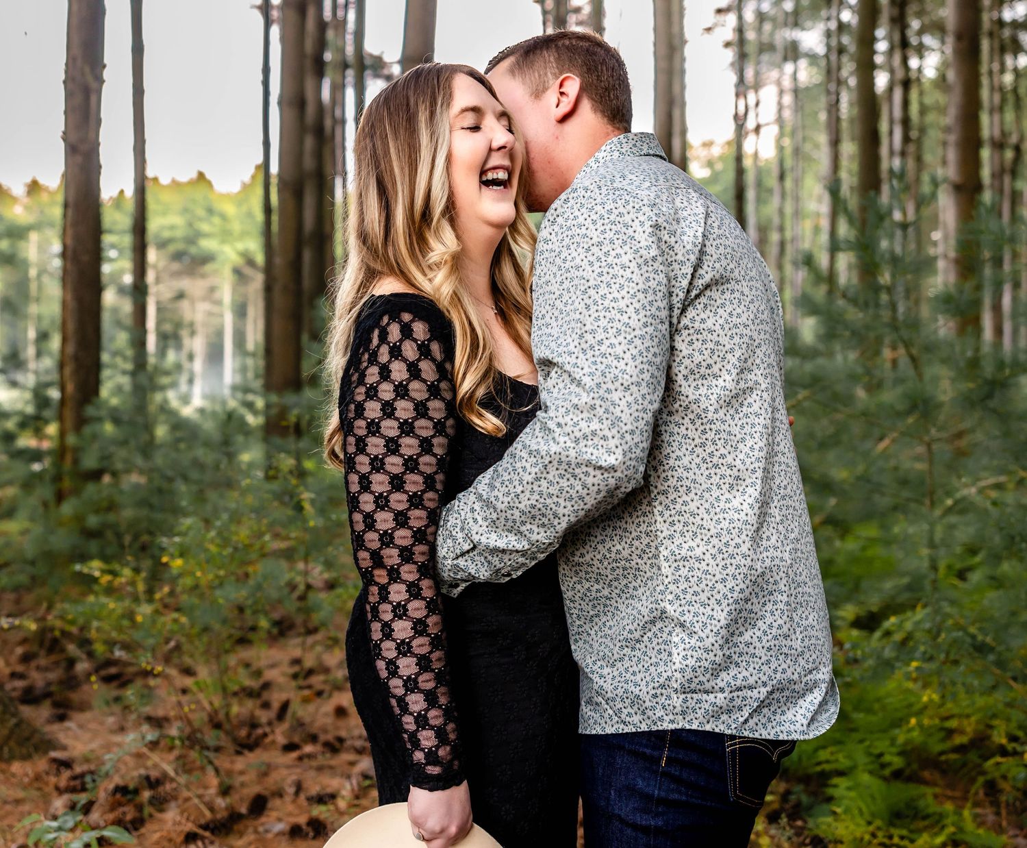 A romantic couple shares a joyful moment in a forest setting wearing casual clothing.
