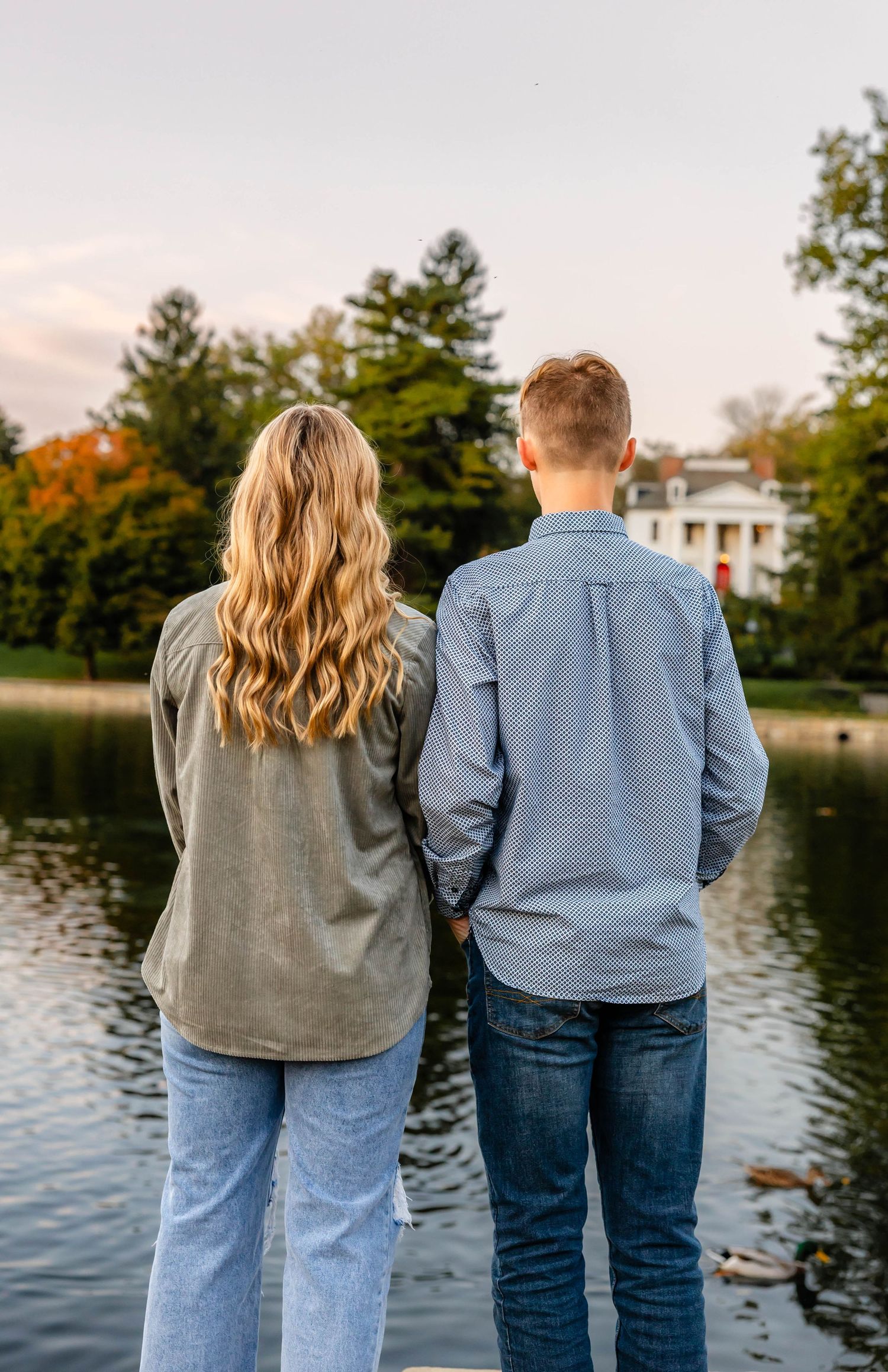 Back view of couple admiring a serene lake with fall foliage and white building in distance.