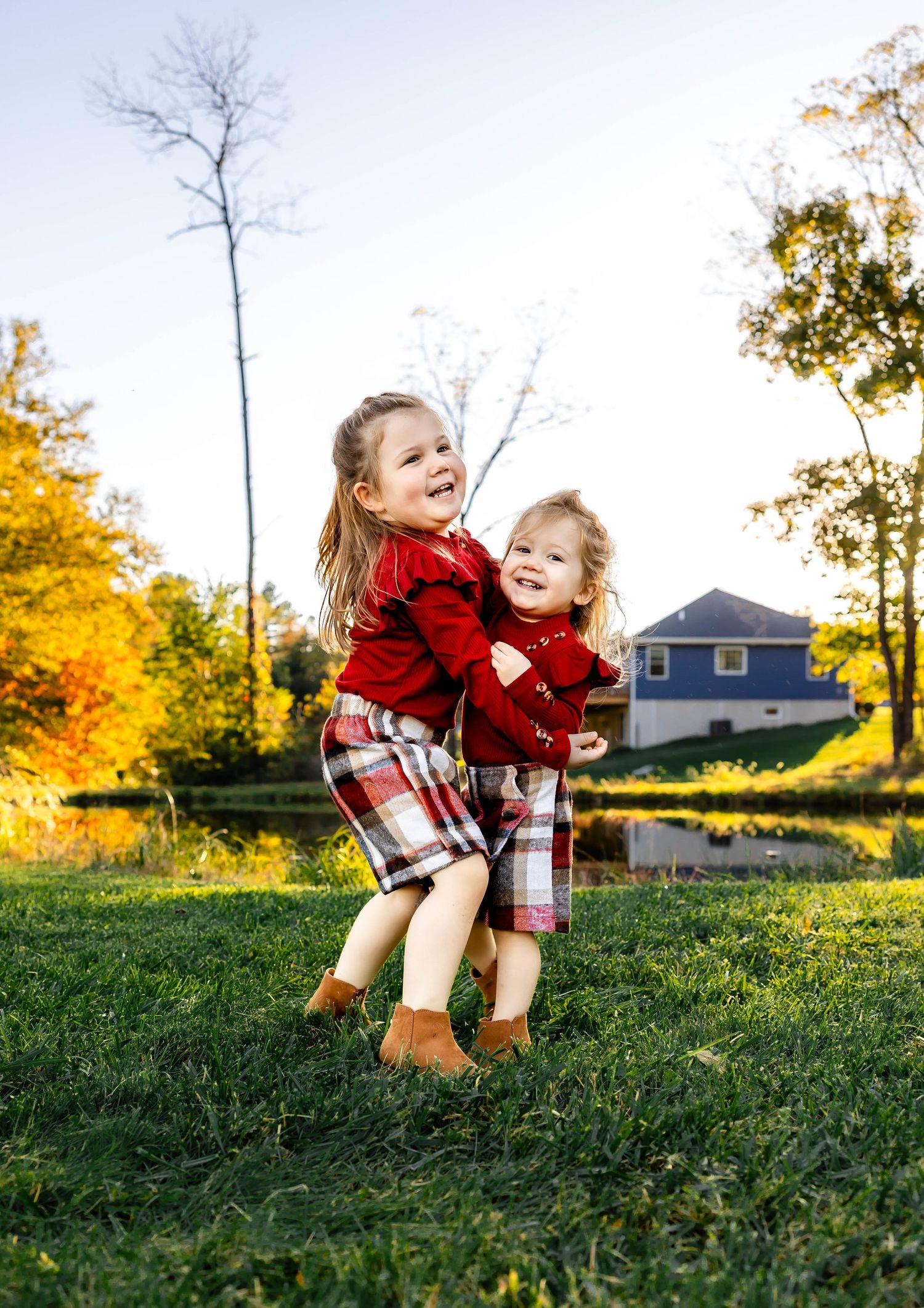Two children in matching red outfits jump together on grass with blue house and fall foliage behind them.