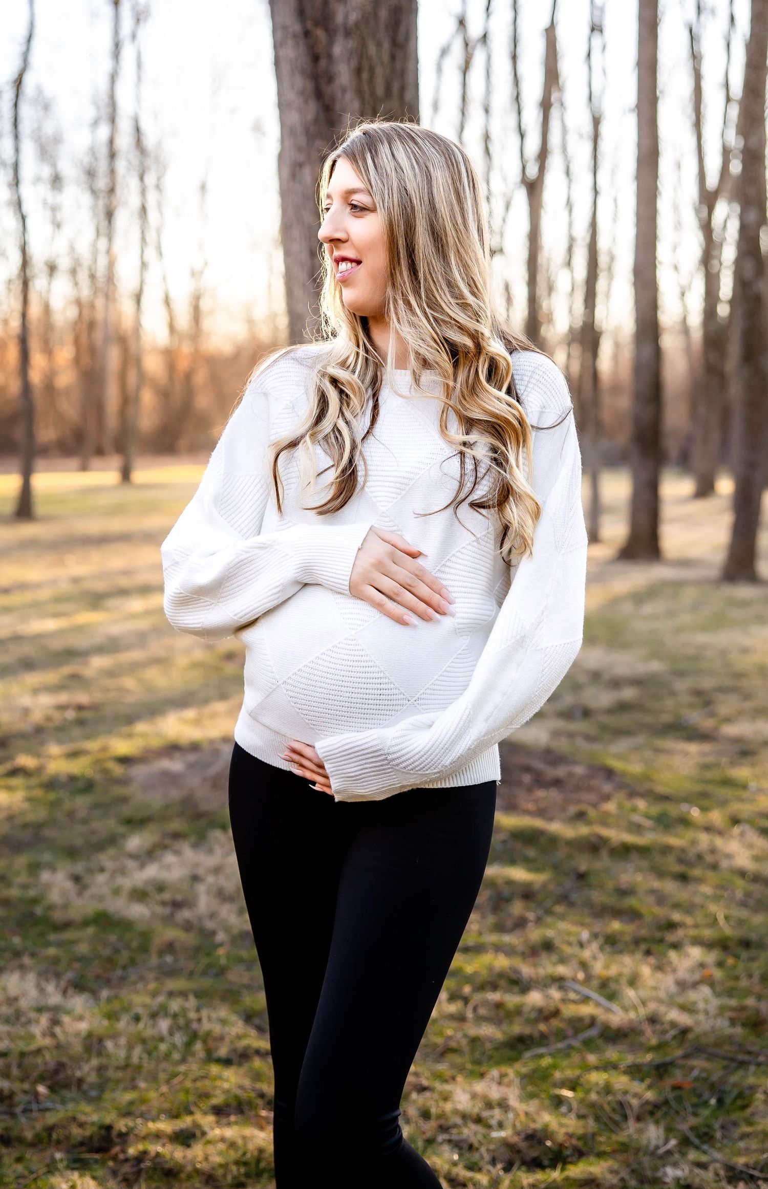 Maternity photo of woman in white sweater standing in wooded setting during golden hour.
