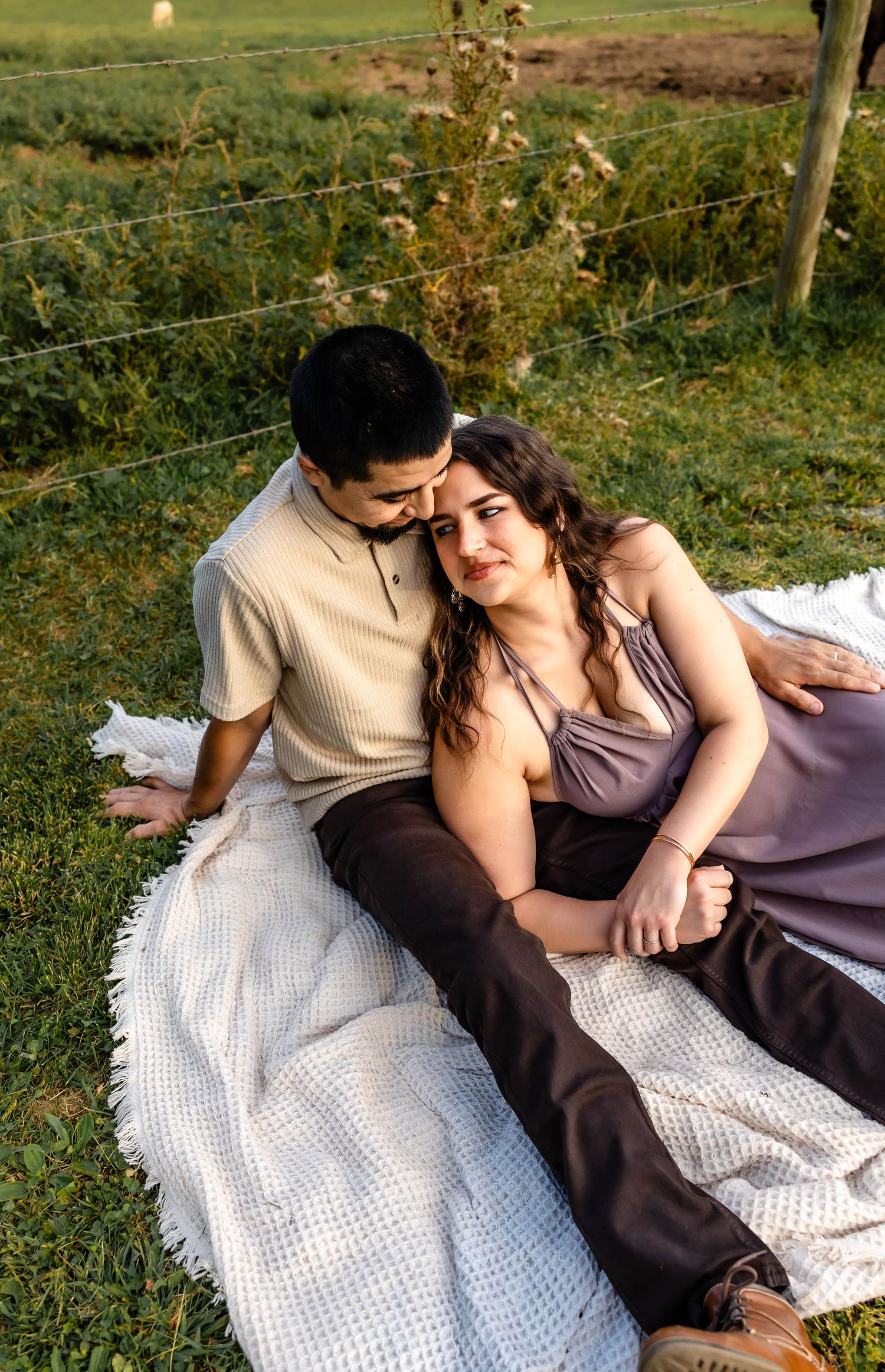 Romantic countryside picnic scene with couple resting on white blanket near wire fence.