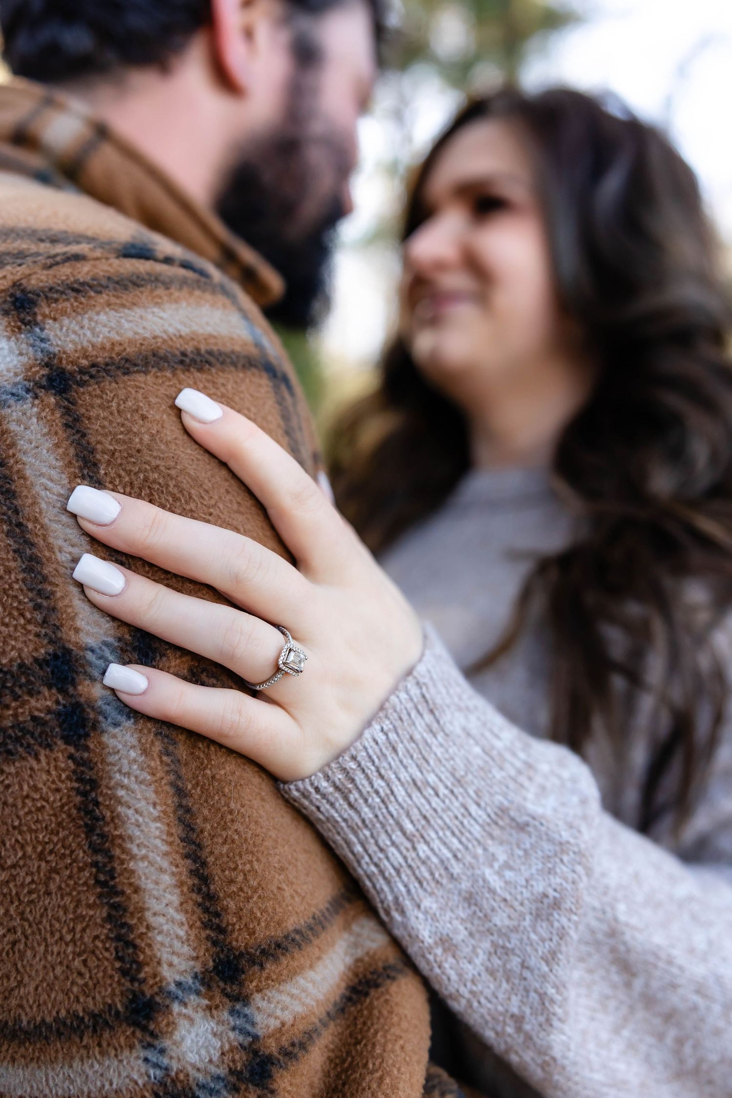 Close-up of engagement ring while couple embraces in forest setting.