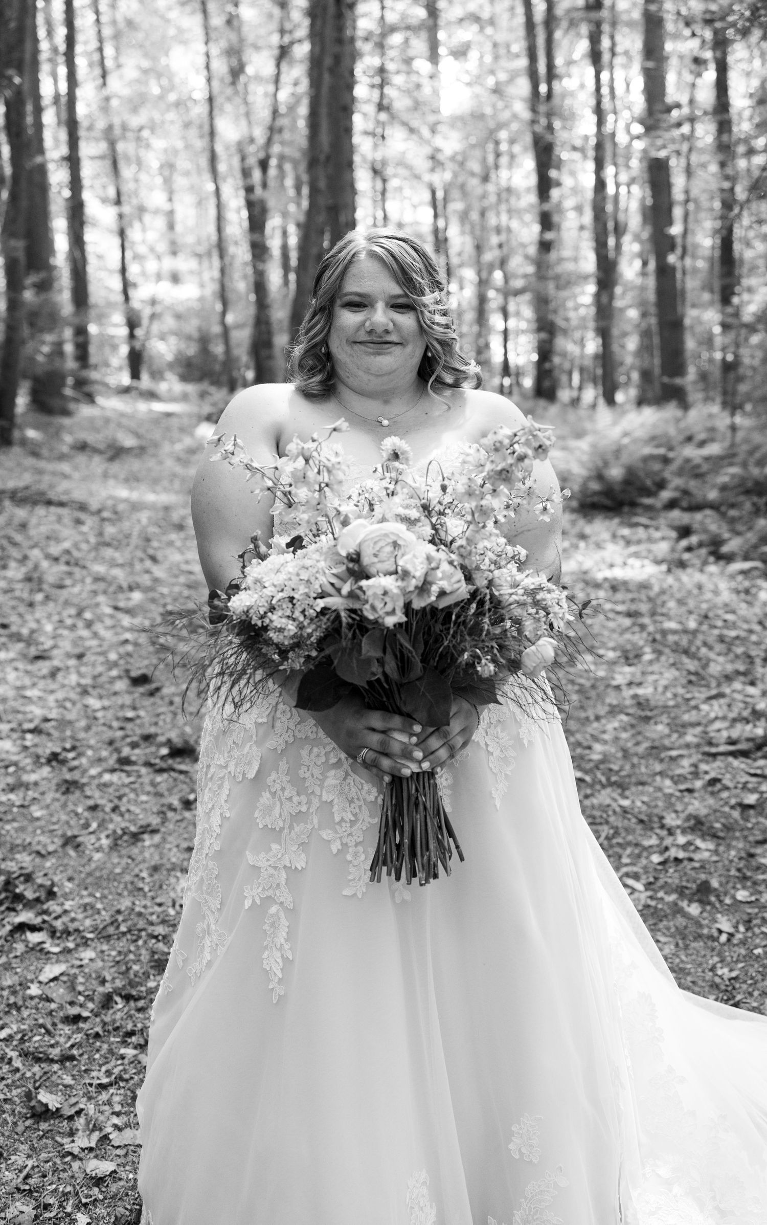 A bride in a lace wedding dress holds a gorgeous bouquet in a forest setting.
