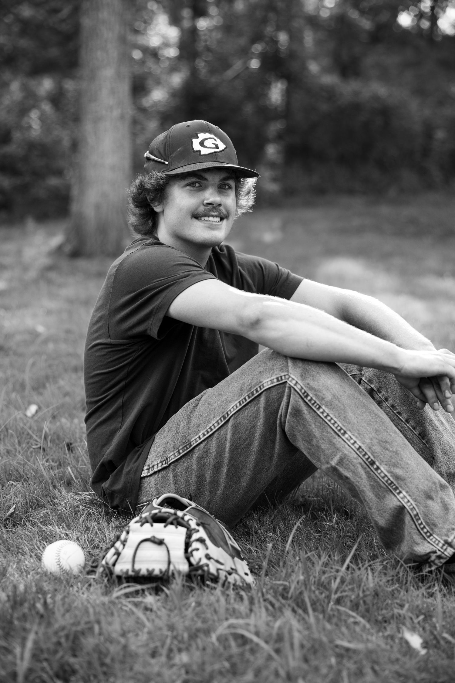 Black and white portrait of baseball player sitting in grass with glove and ball.