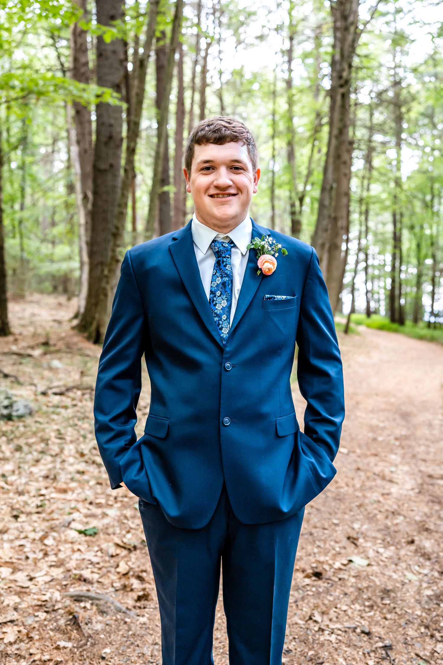 Groom in navy suit with floral tie poses on forest path.